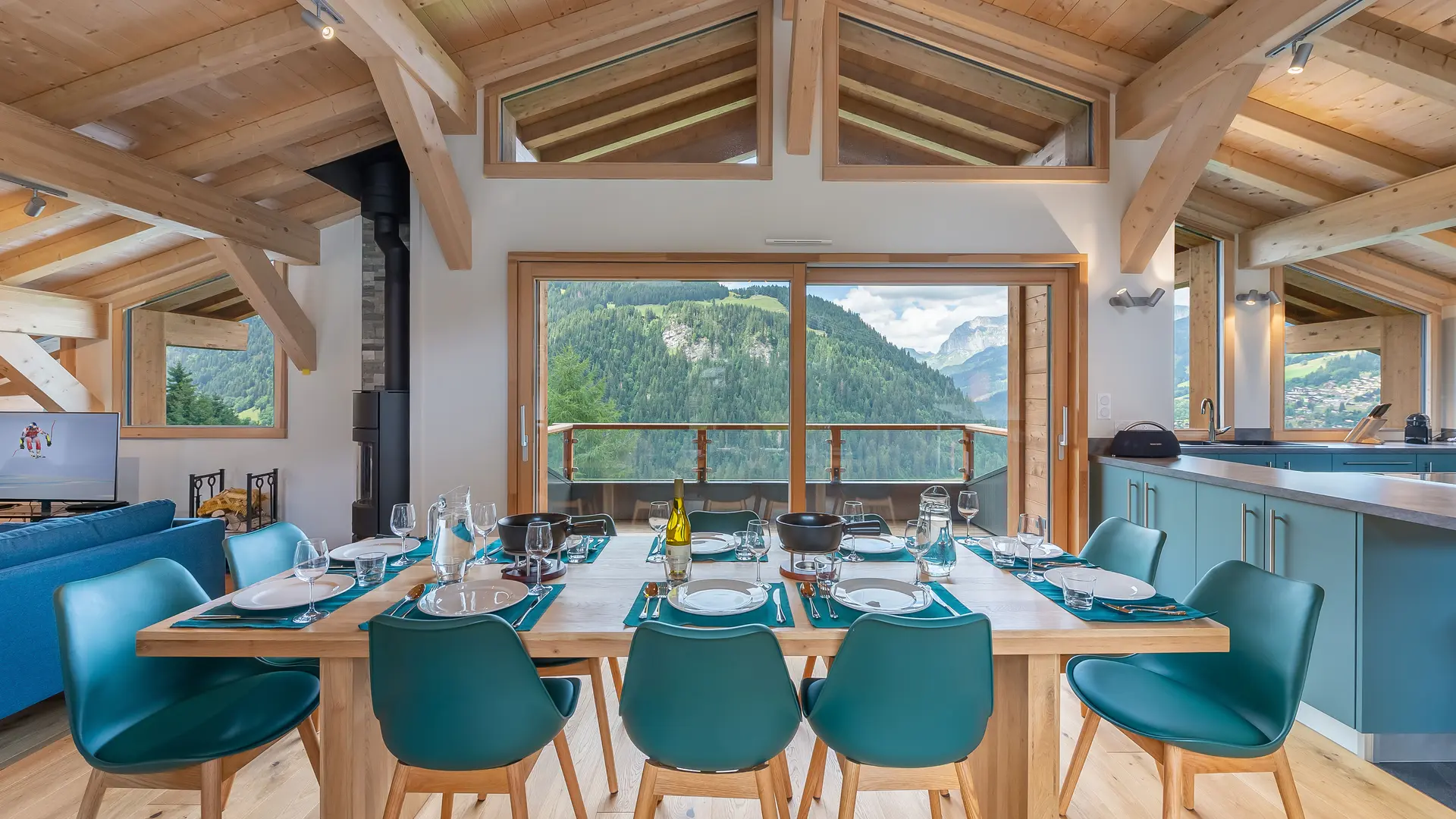 Salle à manger avec sa grande table, chaises bleues, plafond cathédral, poutres apparentes et grandes baies vitrées, vue sur les montagnes