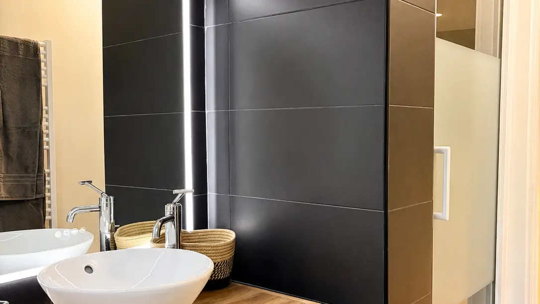 Une salle d'eau moderne comprenant un meuble sous vasque en bois surmonté d'une vasque ronde blanche et d'un miroir, séparé d'une cabine de douche vitrée par une cloison noire.