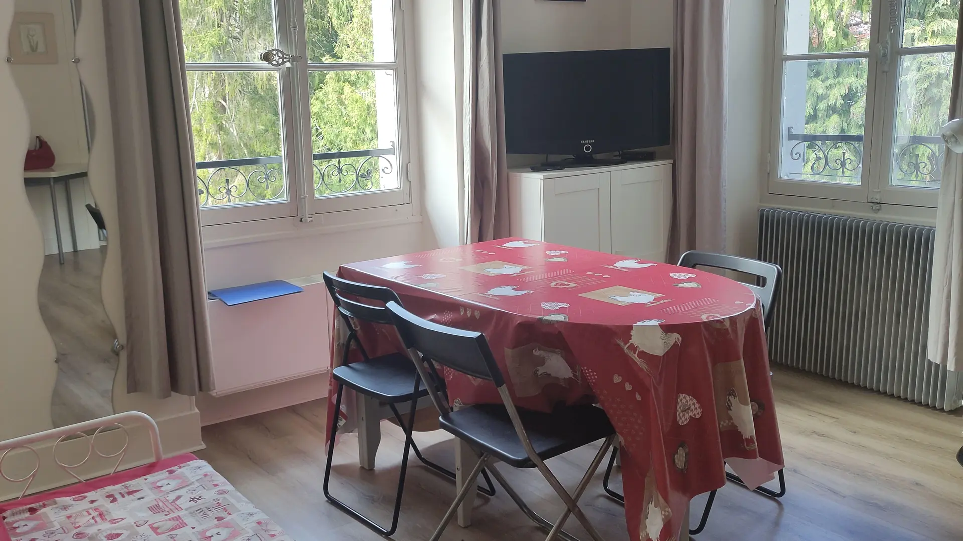 Une grande pièce lumineuse caractérisée par deux hautes fenêtres encadrées de rideaux sombres, meublée au centre d'une longue table recouverte d'une nappe rouge entourée de chaises sombres, et possédant un tapis à motifs au premier plan.