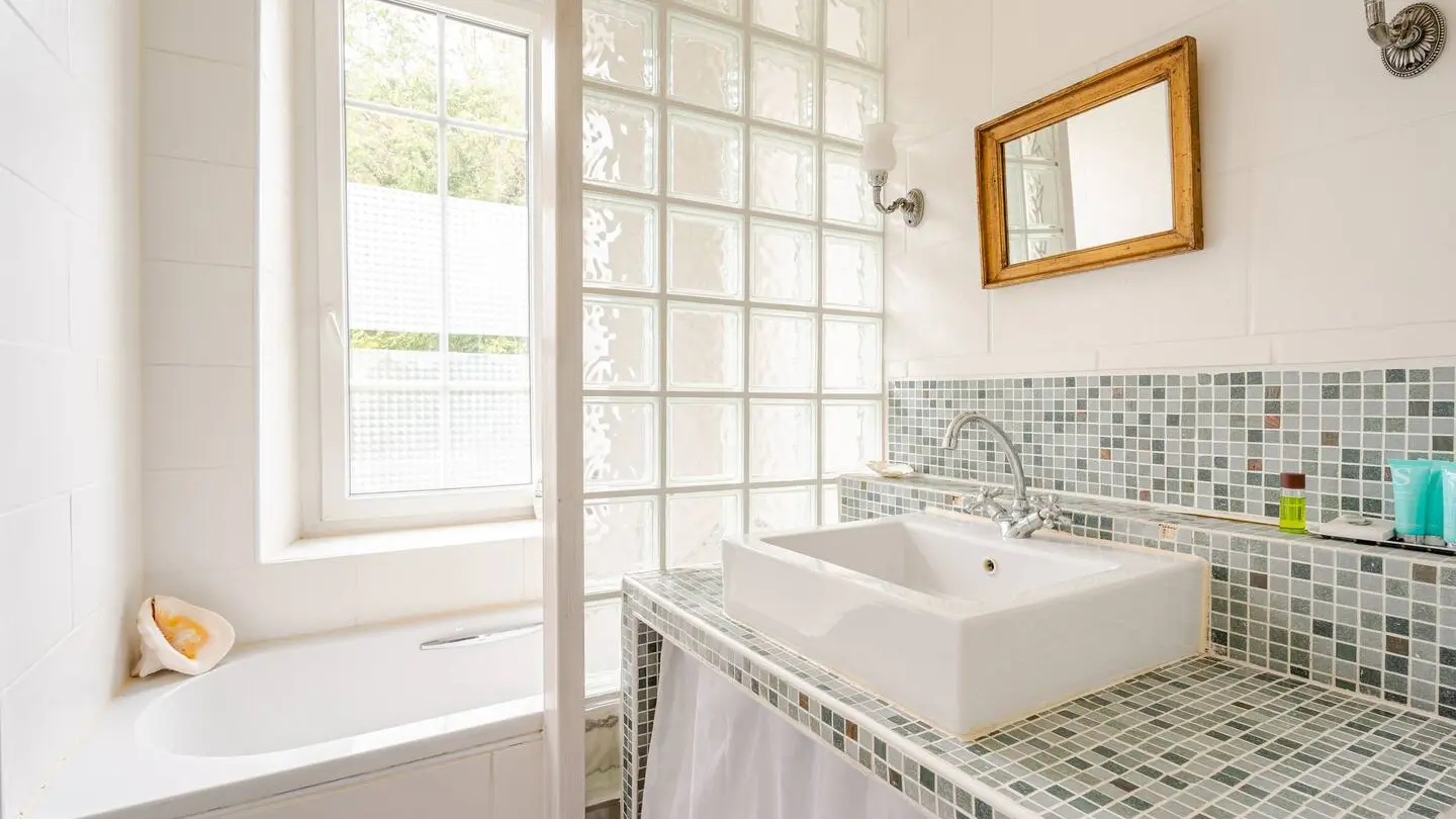 Cette salle de bains est équipée d'une baignoire, d'un lavabo posé sur un meuble, d'une douche séparée par une cloison en verre et d'un miroir. Les murs sont carrelés et une fenêtre apporte de la lumière naturelle.