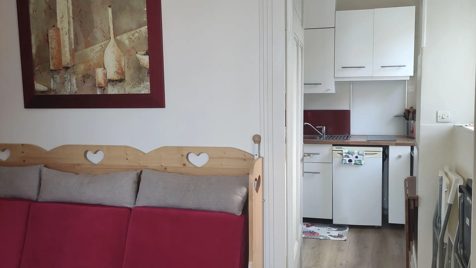 Une pièce à vivre lumineuse meublée d'un canapé rouge et d'un tableau accroché au mur, avec une ouverture donnant sur une cuisine blanche.