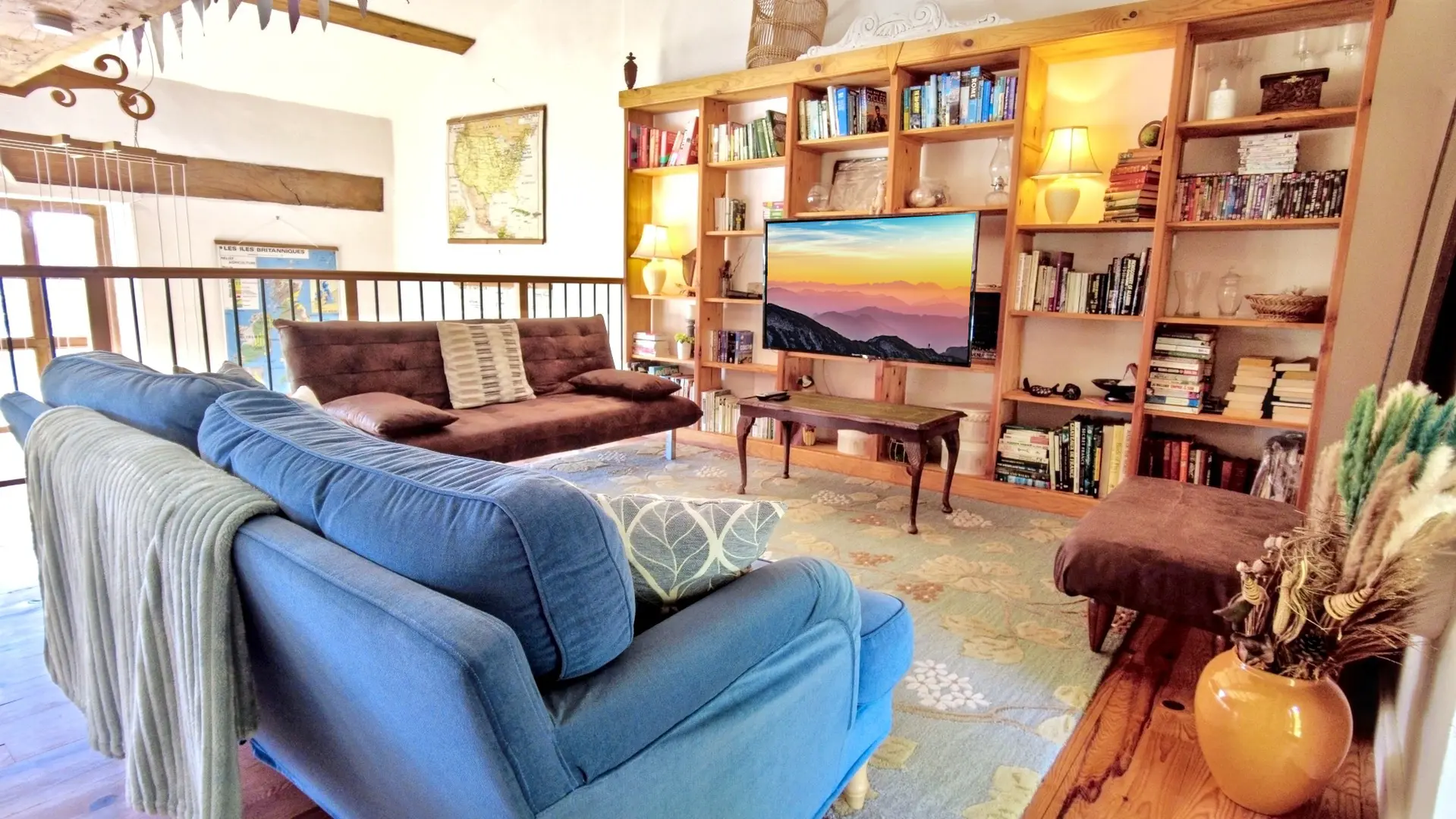 Un salon lumineux et accueillant, doté de planchers en bois. Deux canapés confortables sont orientés vers le centre : un canapé-lit marron foncé texturé sur la gauche, et un fauteuil ou un canapé bleu vif sur la droite, avec des coussins à motifs blancs et bleus. Entre les deux, un tapis à motifs floraux beiges et bleus couvre le sol. Au centre, une petite table basse en bois sombre de style antique est visible. Sur le mur du fond, de grandes étagères encastrées en bois clair remplies de livres et d'objets décoratifs entourent un écran de télévision plat qui affiche un paysage montagneux au lever ou au coucher du soleil. À droite, un grand miroir sur pied au cadre sombre reflète le reste de la pièce. La pièce présente des poutres en bois apparentes au plafond et une fenêtre à droite qui laisse entrer la lumière naturelle.