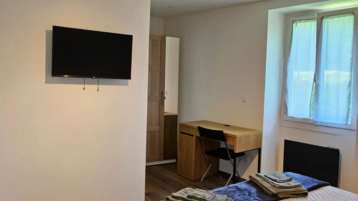 La chambre est équipée d'un grand lit, d'un bureau, d'une télévision fixée au mur et d'une fenêtre. Le sol est en bois et le plafond est traversé par une poutre en bois