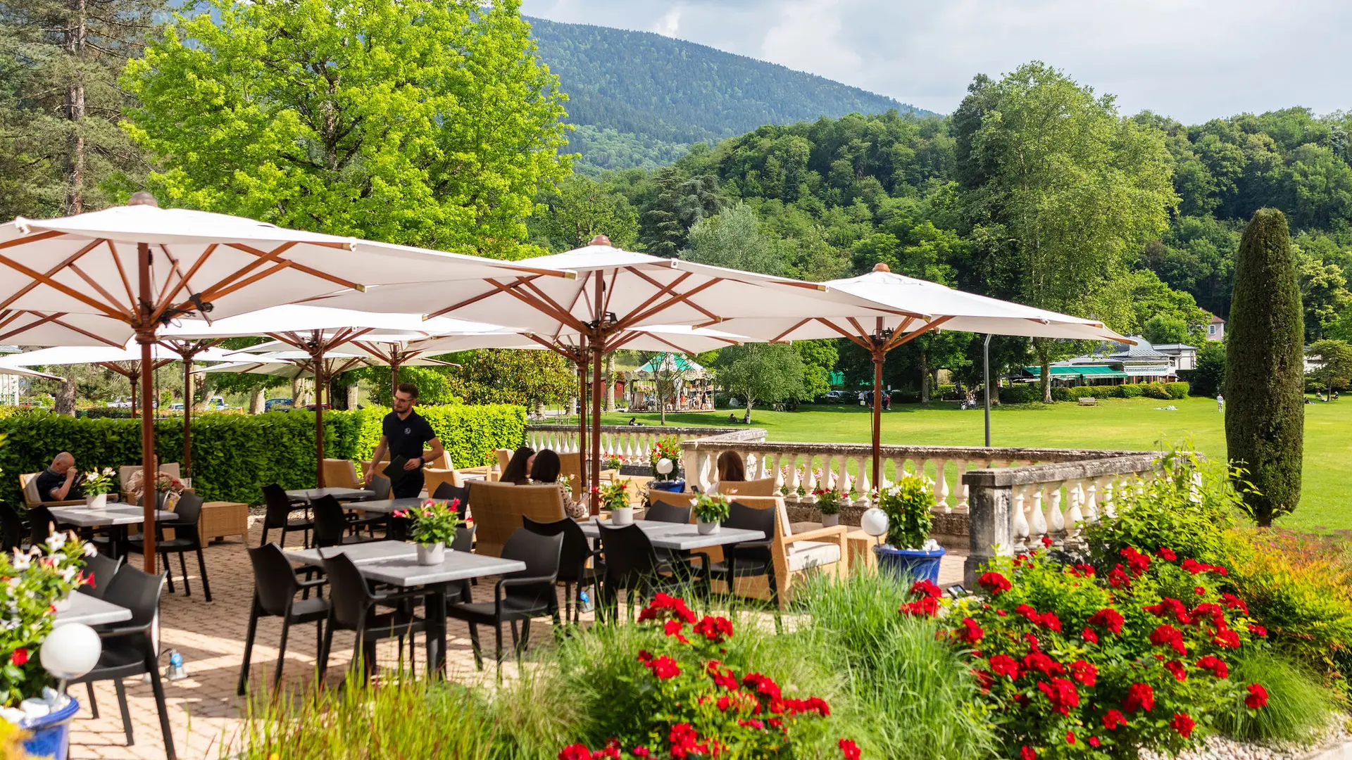 Cette terrasse en plein air comprend plusieurs tables et chaises disposées sous de grands parasols, avec une vue sur le parc thermal d'Uriage.