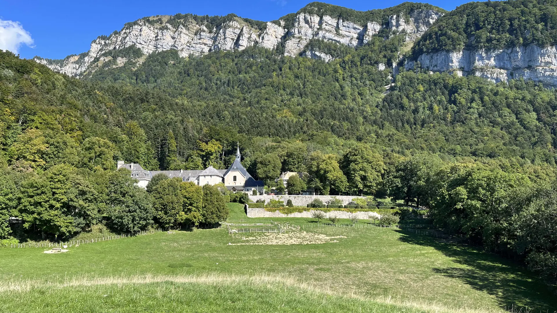 Splendide vue du Monastère, fondu dans son environnement verdoyant où la nature est omniprésente. Au premier plan, une prairie amène le regard jusqu'à l'édifice religieux, au pied de la montagne sous ciel bleu