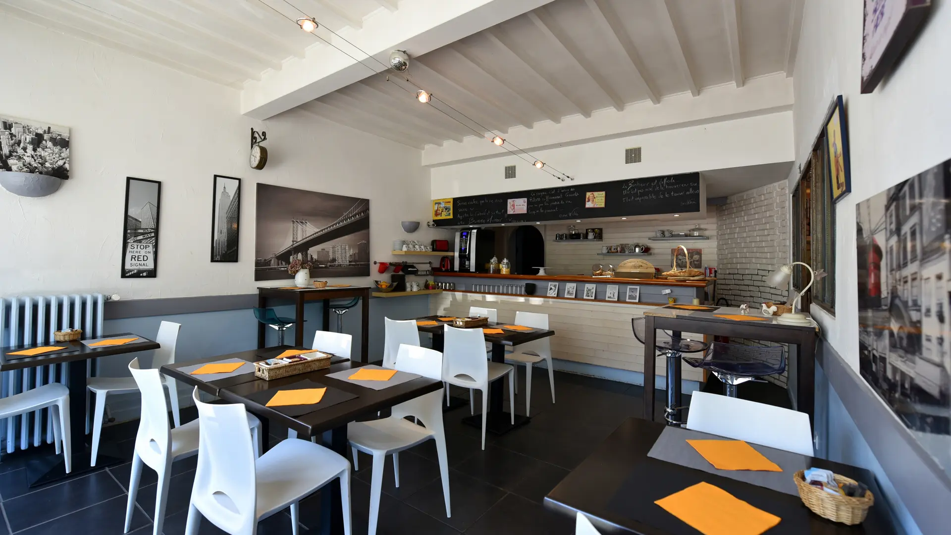 Salle de petit déjeuner avec six tables dressées dont deux tables hautes pour deux personnes. Un buffet petit déjeuner est installé en arrière plan. Les murs sont décorés de nombreux cadres et d’une horloge
