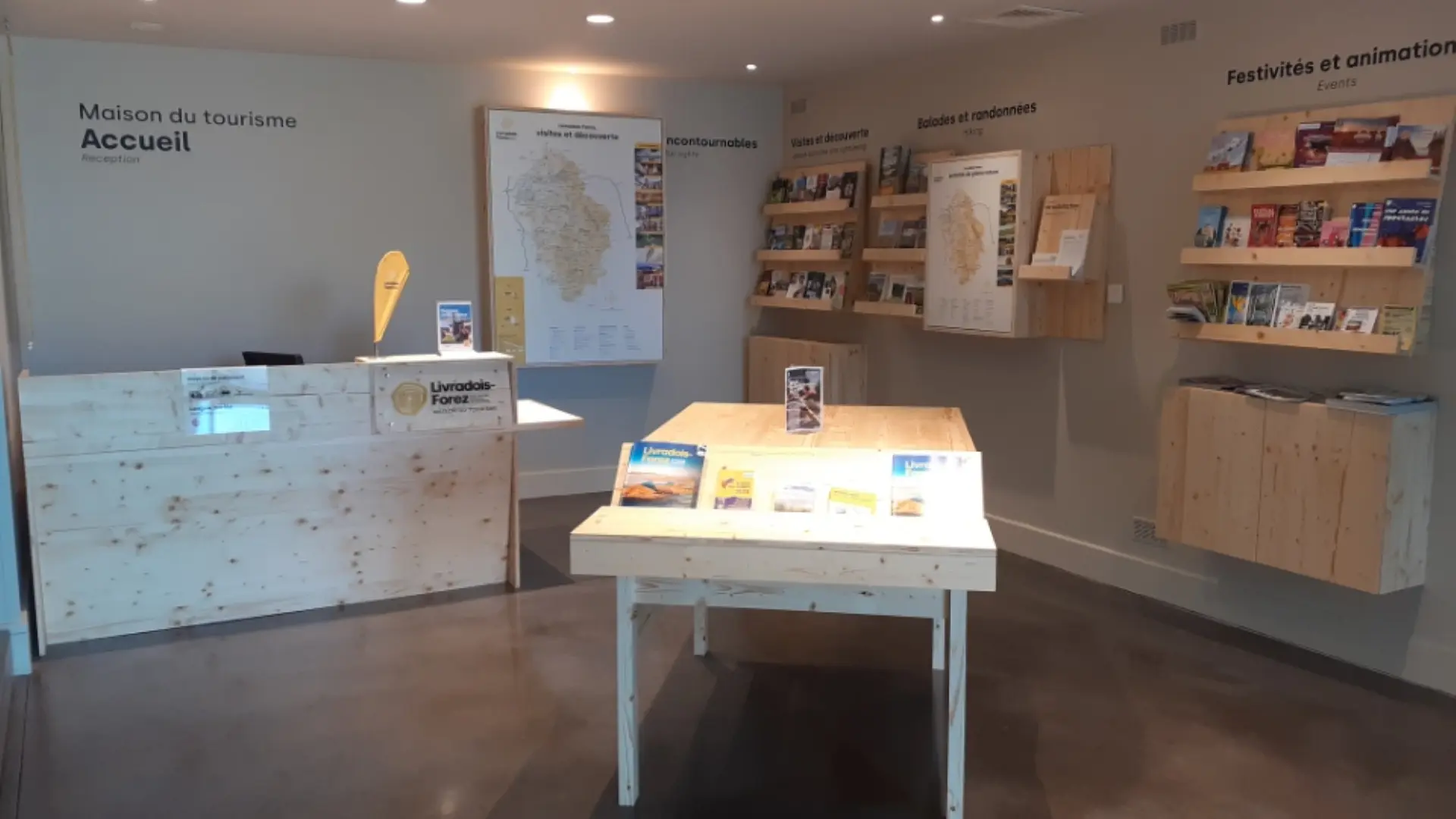 Intérieur du Point d'information touristique avec meubles de présentation pour la documentation du territoire. Au centre se trouve le piano pour échanger avec le conseiller en séjour. L'espace est ouvert et accueillant avec du mobilier en bois de la région Livradois-Forez