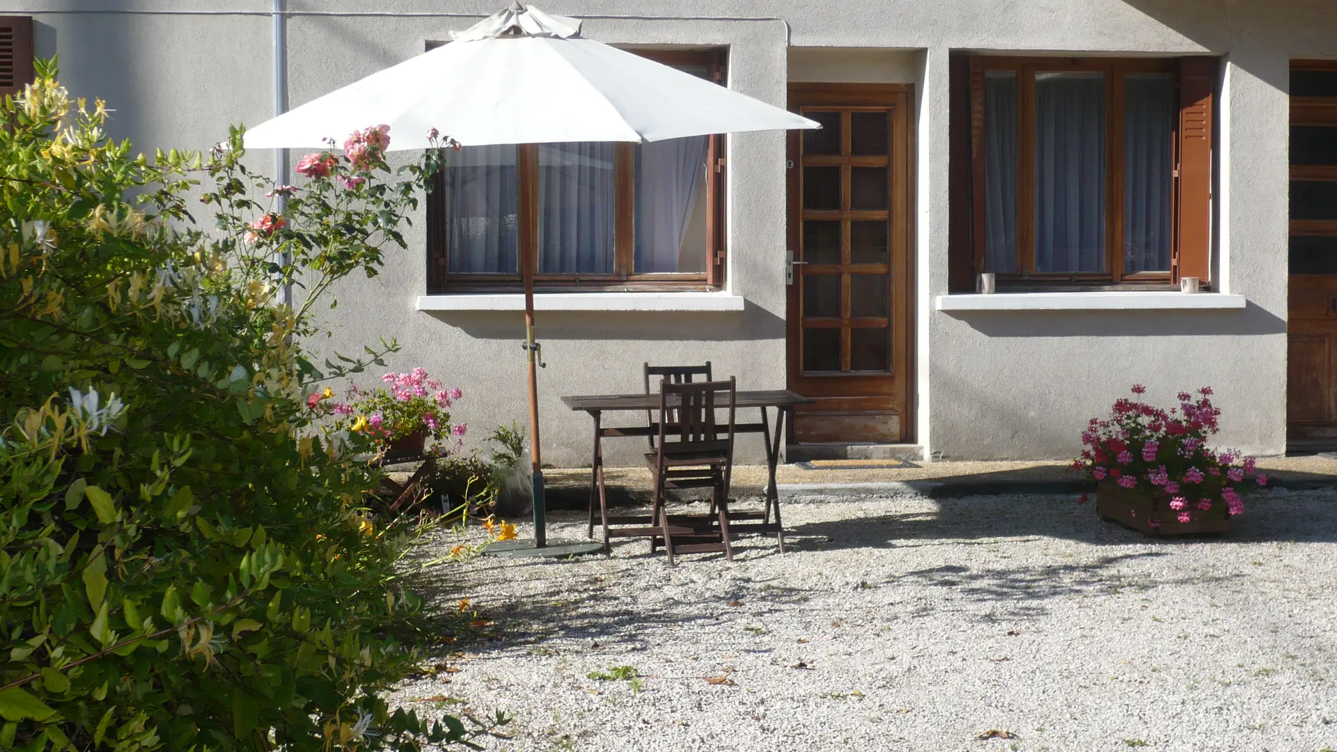 La terrasse, adjacente à la maison, se compose d'un sol en gravier et est aménagée avec un salon de jardin en bois sous un parasol blanc.