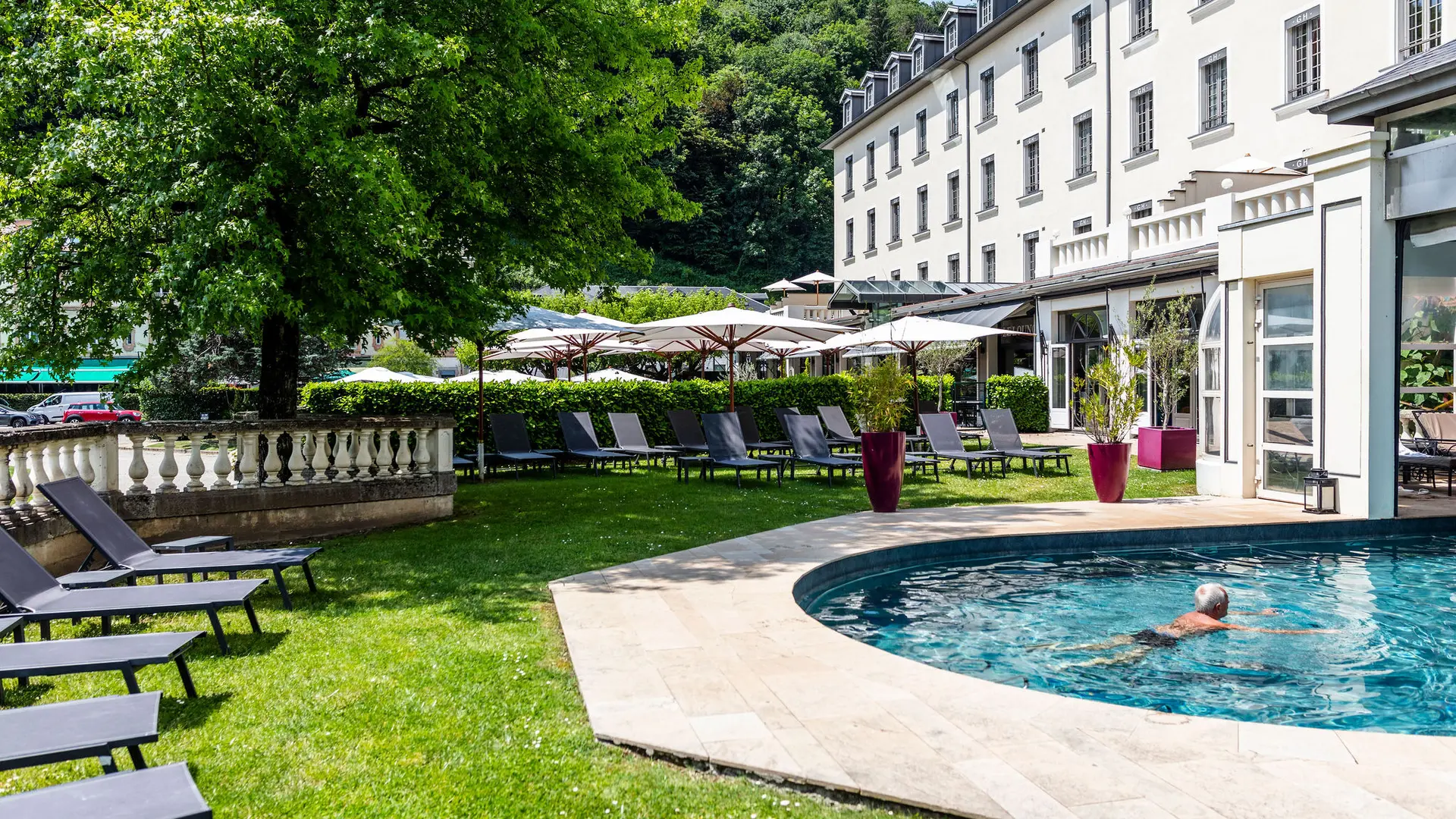 Une piscine extérieure-intérieure avec un homme se baignant. La piscine est entourée par un espace engazonné avec des transats gris. On aperçoit le grand hôtel et sa terrasse au fond.