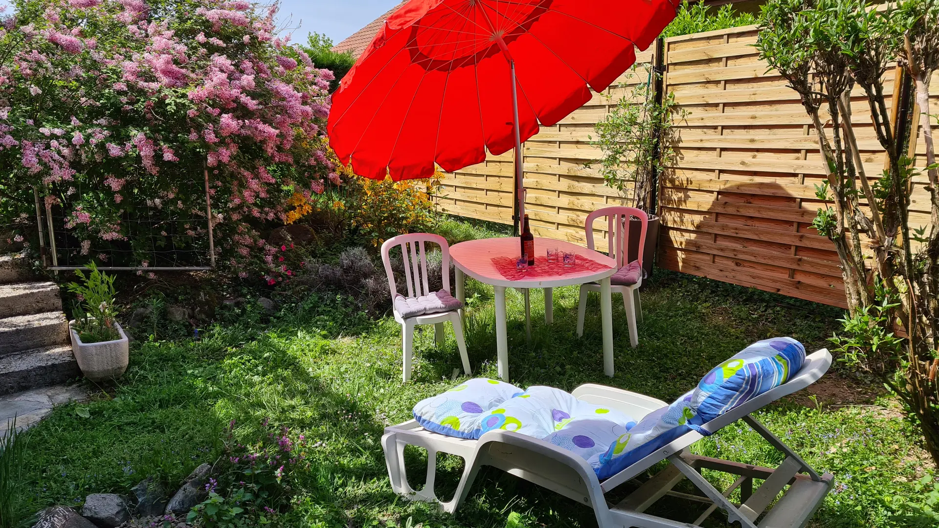 Le jardin est équipé d'une table de jardin et de chaises, ainsi que de chaises longues. Un parasol rouge offre de l'ombre. La végétation est composée principalement de buissons et d'arbustes.