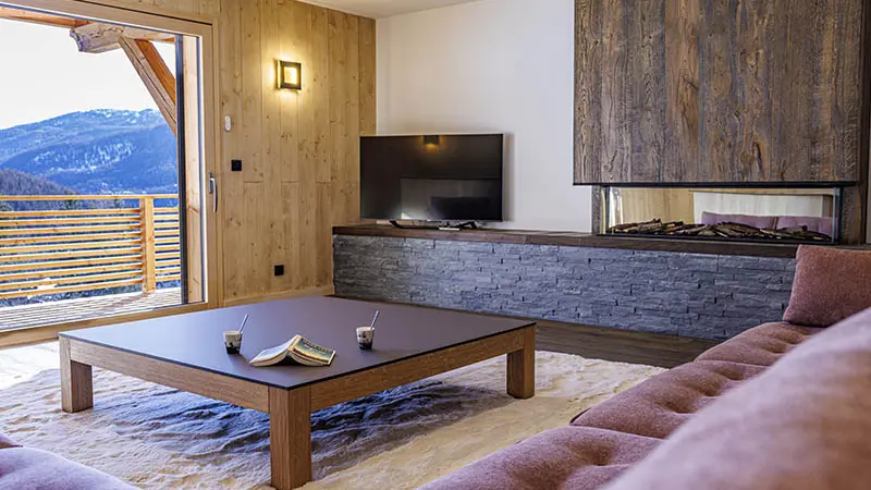 Grand et lumineux séjour, table basse, canapé, TV, ameublement bois et pierre, porte-fenêtre donnant sur un balcon en bois et paysage enneigé