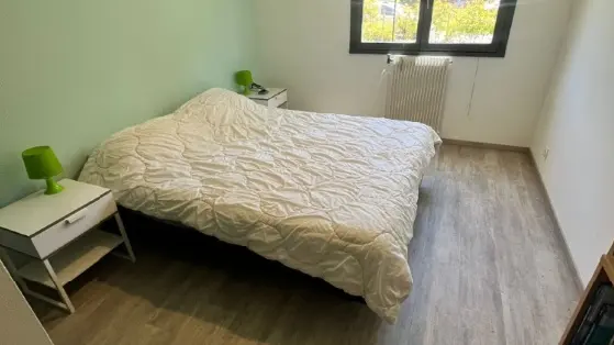 Une chambre avec des murs peints en blanc et vert, un lit double au centre avec une couette blanche, et une fenêtre qui laisse passer la lumière du jour.
