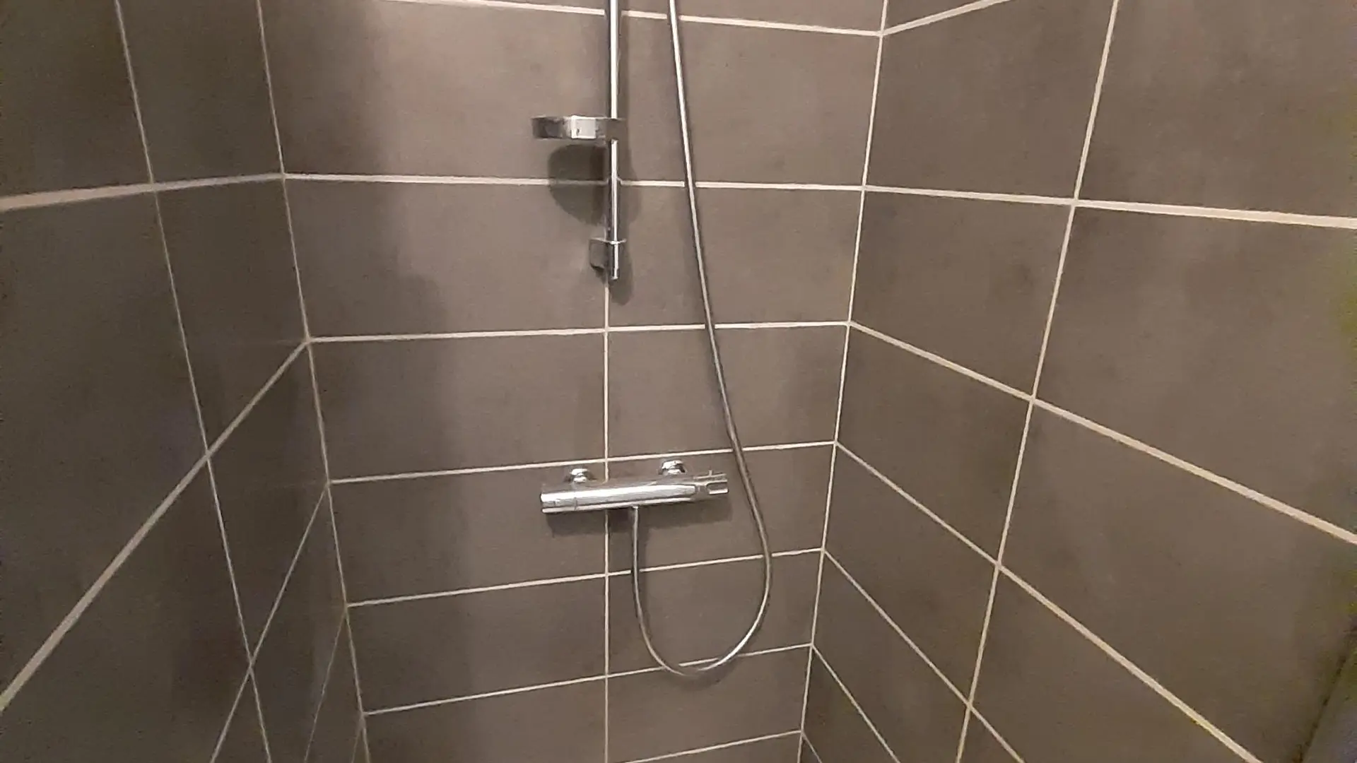 Une salle d'eau avec une douche. Les murs sont carrelés. ne étagère en verre, fixée au mur, permet de poser les produits de douche.