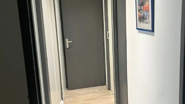 Un couloir avec un sol en parquet clair, des murs blancs et plusieurs portes sombres sur les côtés, une armoire avec des portes coulissantes blanches, et un cadre accroché au mur du fond.