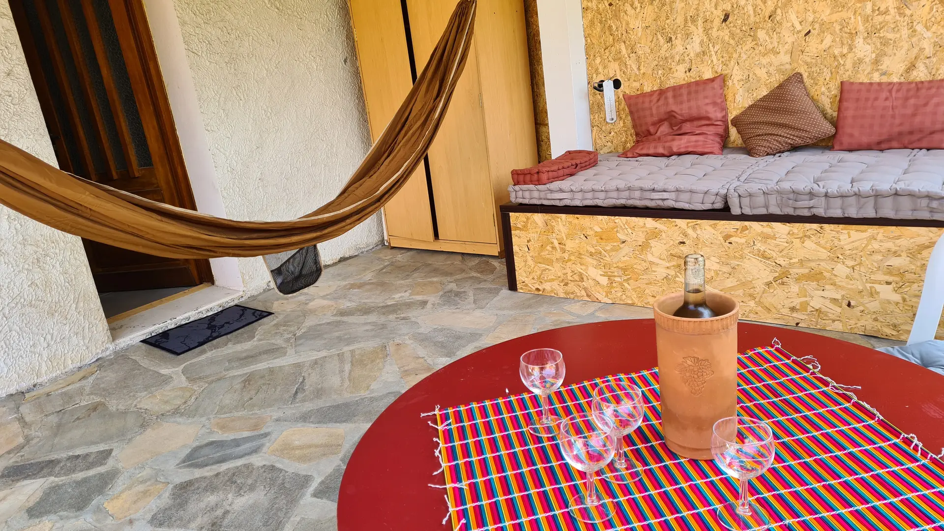 La terrasse, couverte et ombragée, est aménagée avec un hamac suspendu, un banc moelleux recouvert de coussins et une table basse ronde sur laquelle se trouvent une bouteille et des verres.