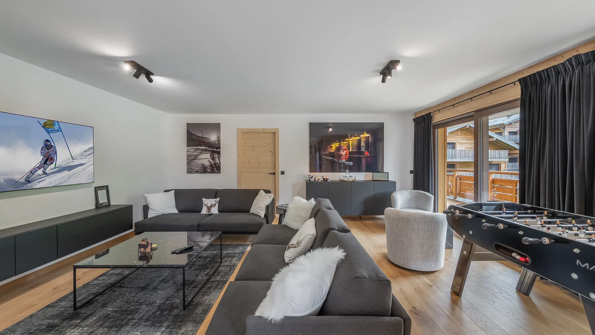 Espace salon avec grands canapés, table basse, télévision, baby foot, accès au balcon par grande baie vitrée