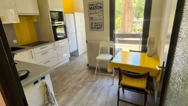 Une cuisine avec un sol en parquet clair, des placards de cuisine blancs, des murs jaunes et une table avec une nappe jaune près d'une fenêtre.