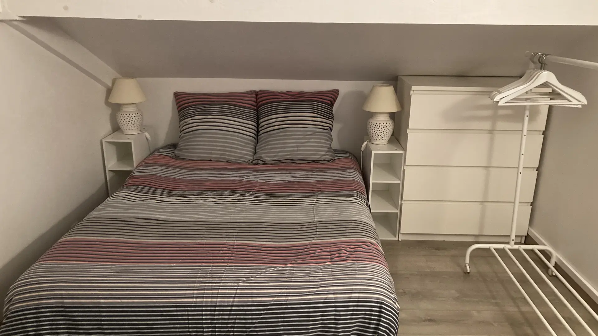 C'est une chambre mansardée, éclairée, contenant un lit double fait d'une couette rayée, encadré par deux petites tables de chevet avec des lampes, une commode blanche à tiroirs et un portant à vêtements métallique.
