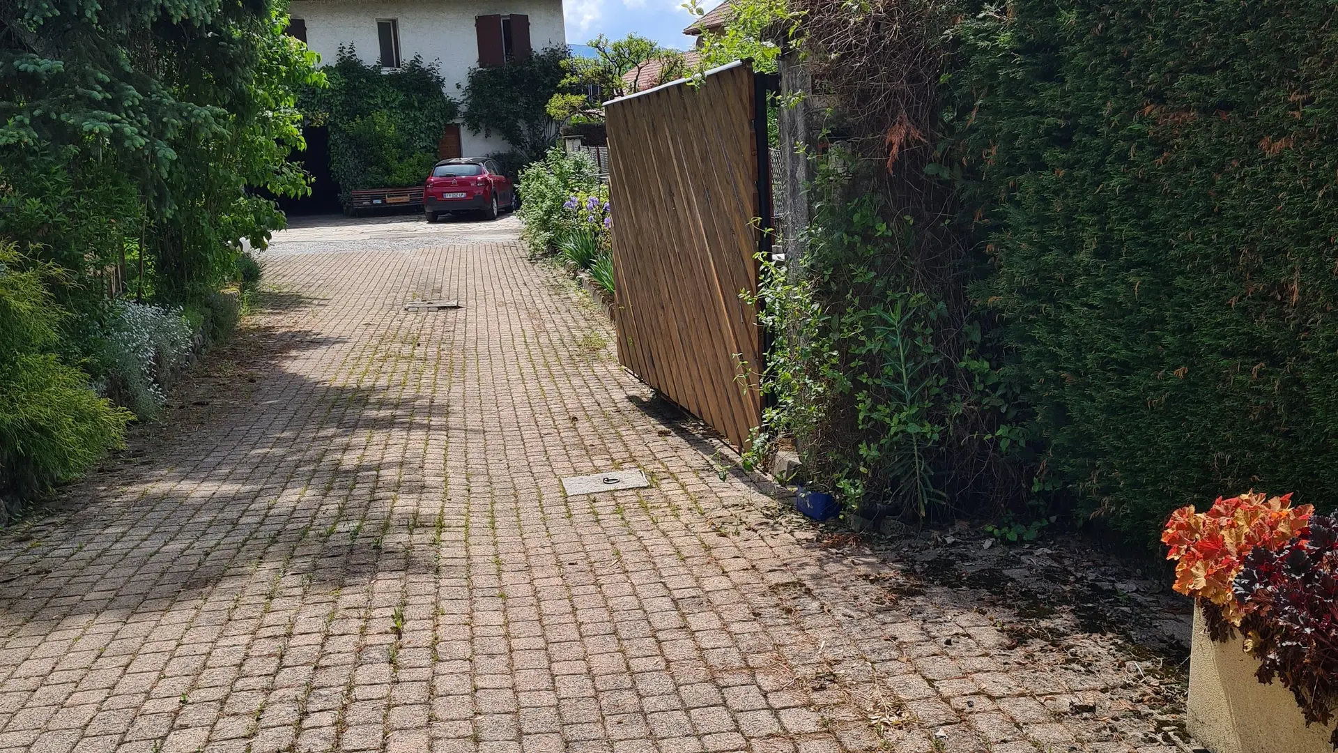 L'allée pavée monte légèrement vers une maison entourée de végétation. Un portail en bois ferme l'accès à la propriété.