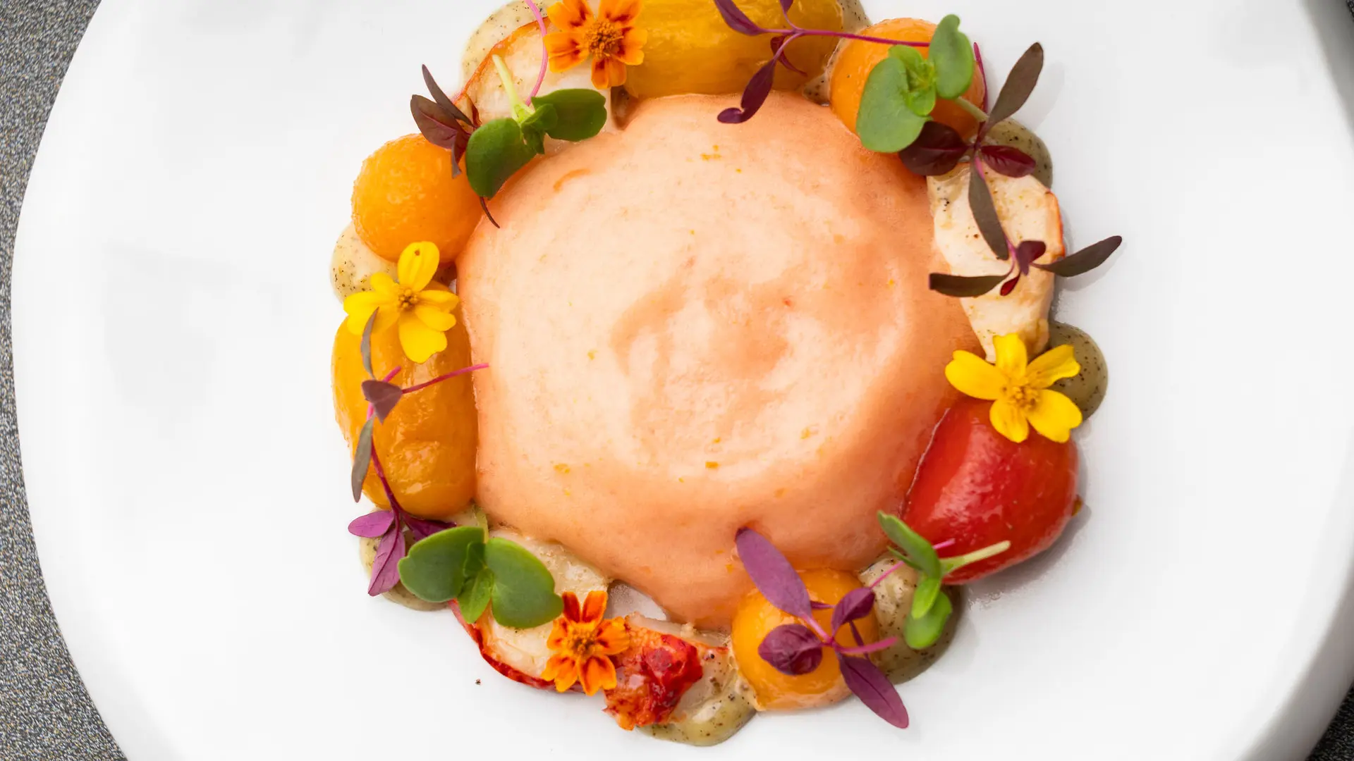 Au centre d'une assiette blanche, un cercle de mousse légère de couleur pêche avec petites feuilles et fleurs comestibles de couleurs vives.