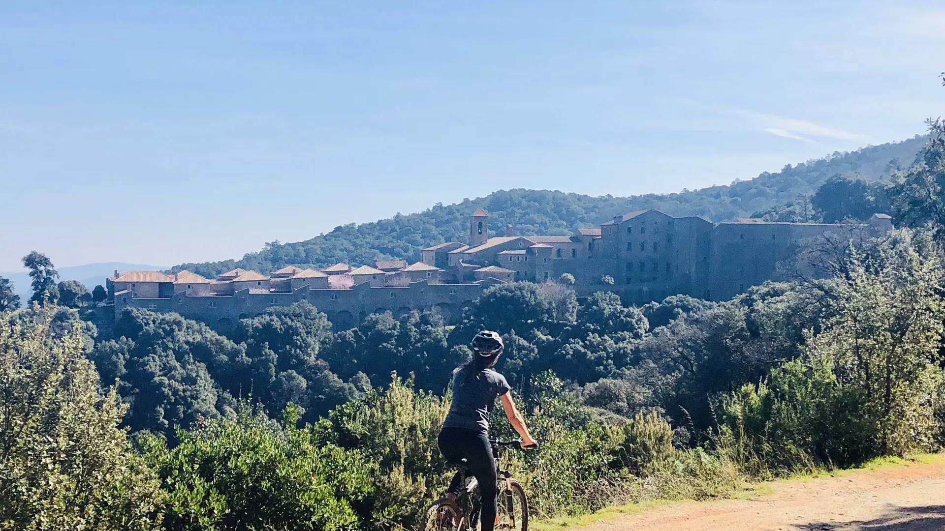 Cycliste sur un sentier en terre avec la vue au loin de la Chartreuse de la Verne posée au milieu de la forêt des Maures