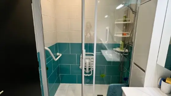 Une salle d'eau, une douche de plain-pied avec un siège blanc rabattable une paroi en verre, des murs carrelés en blanc et turquoise, et un meuble de lavabo blanc sur la droite.