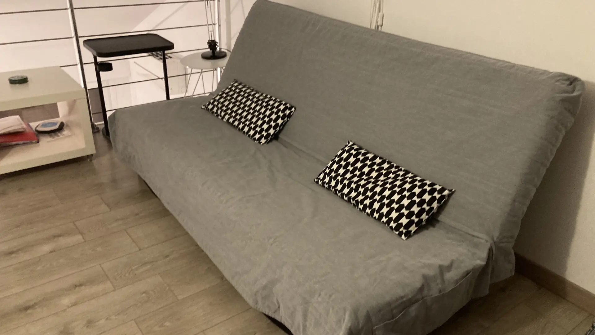 C'est un espace aménagé sous des combles, doté d’un canapé clic-clac gris et à côté duquel se trouvent une petite table d'appoint blanche surmonté d’une lampe de chevet.