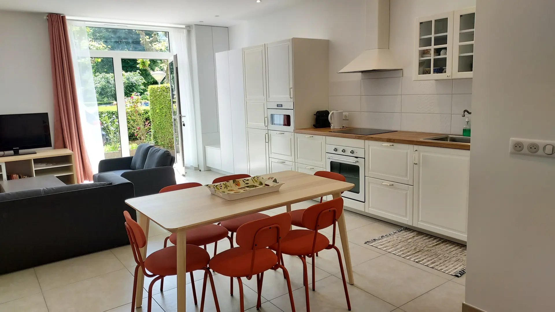 Une pièce de vie ouverte et lumineuse avec une cuisine équipée avec des armoires blanches, un plan de travail et des électroménagers. Un salon attenant est équipé d'un canapé et d'une télévision. Une table en bois avec des chaises rouges est placée entre les deux espaces, créant ainsi un coin repas convivial.