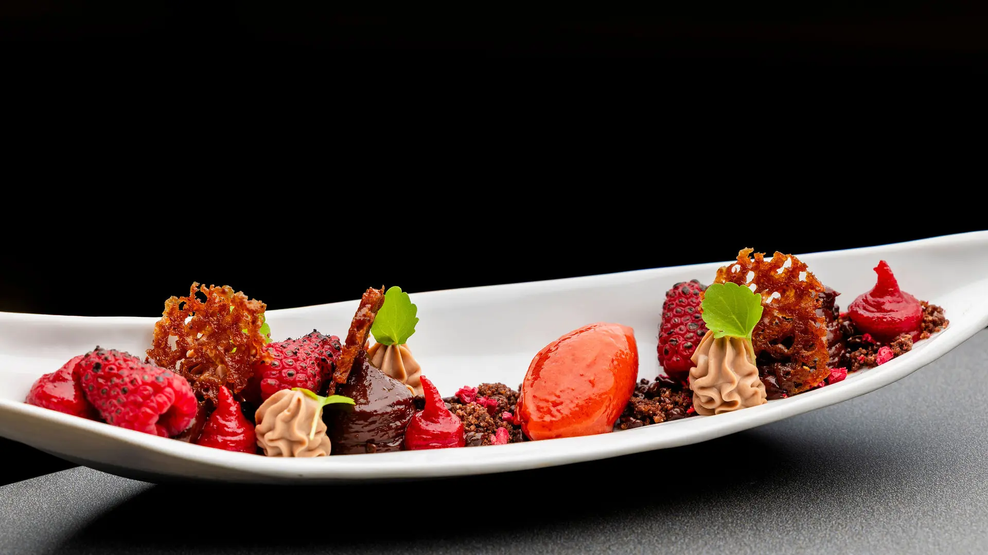 Un dessert artistique est présenté sur une longue assiette blanche incurvée, composé de plusieurs éléments dont une quenelle rouge vif, des framboises entières, des pointes de crème légère, des morceaux de tuile croustillante et des miettes foncées.