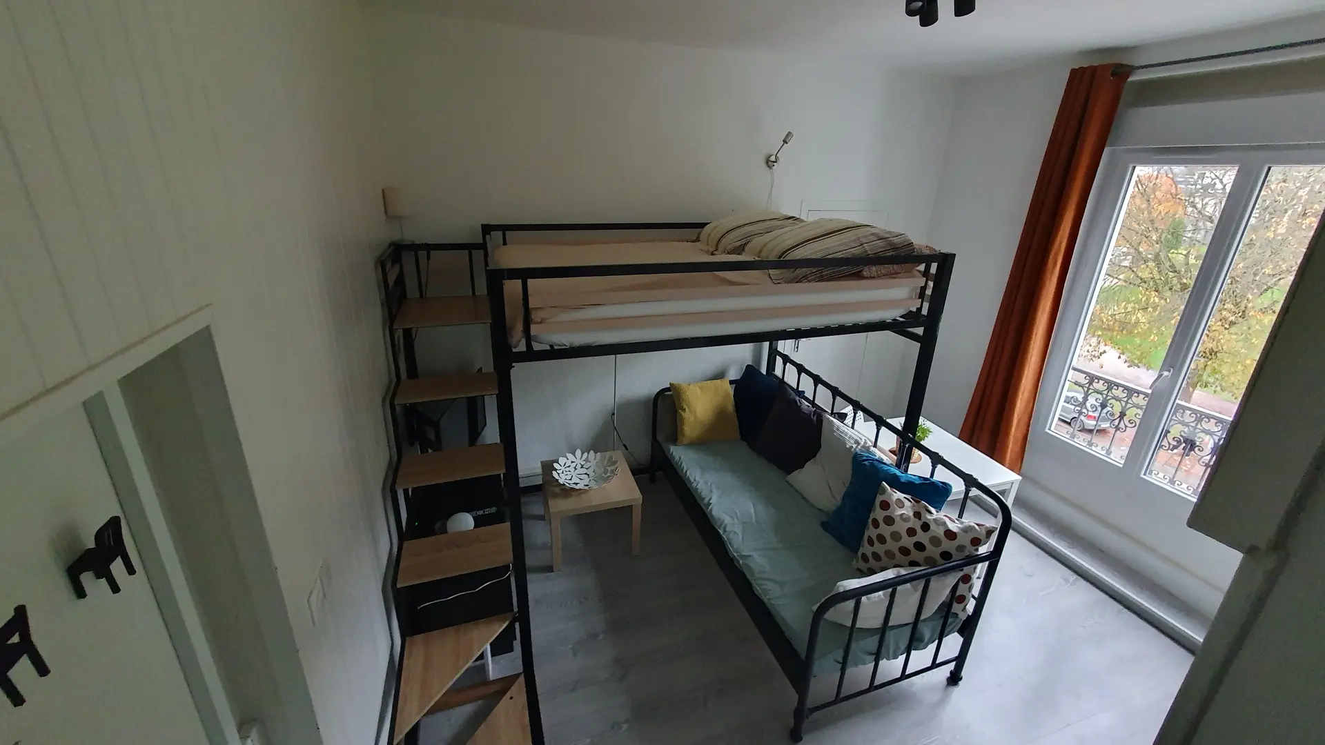 La pièce est un studio de style moderne vue d'en haut, dominé par un grand lit mezzanine en métal noir avec une couchette inférieure utilisée comme canapé, une étagère latérale servant d'escalier, et une porte-fenêtre à droite bordée d'un rideau orange.