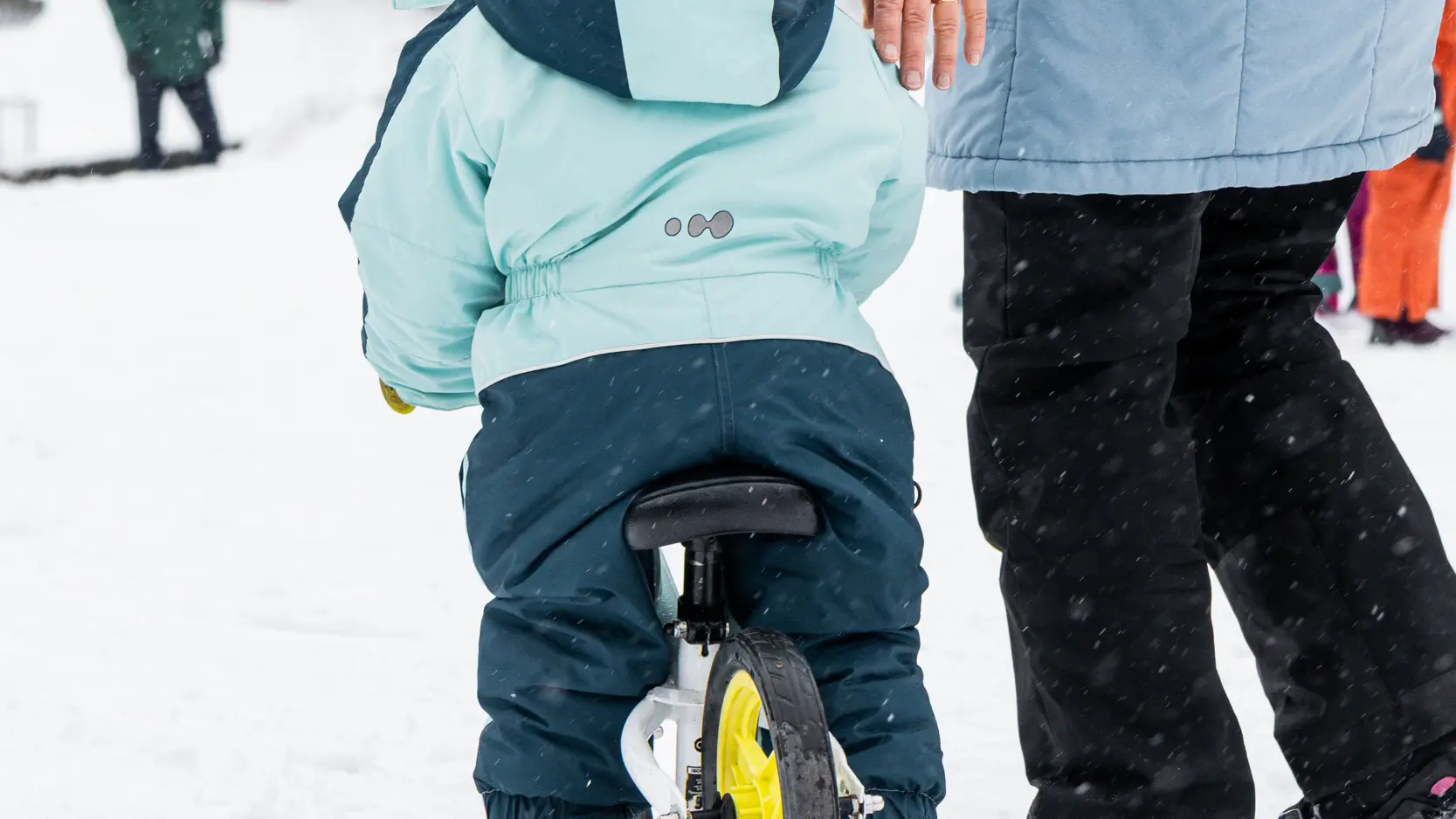 Un enfant de dos, portant un casque et une combinaison de ski bleu clair, est assis sur un petit vélo-ski blanc. Un adulte marche à ses côtés pour l'accompagner sur la neige.