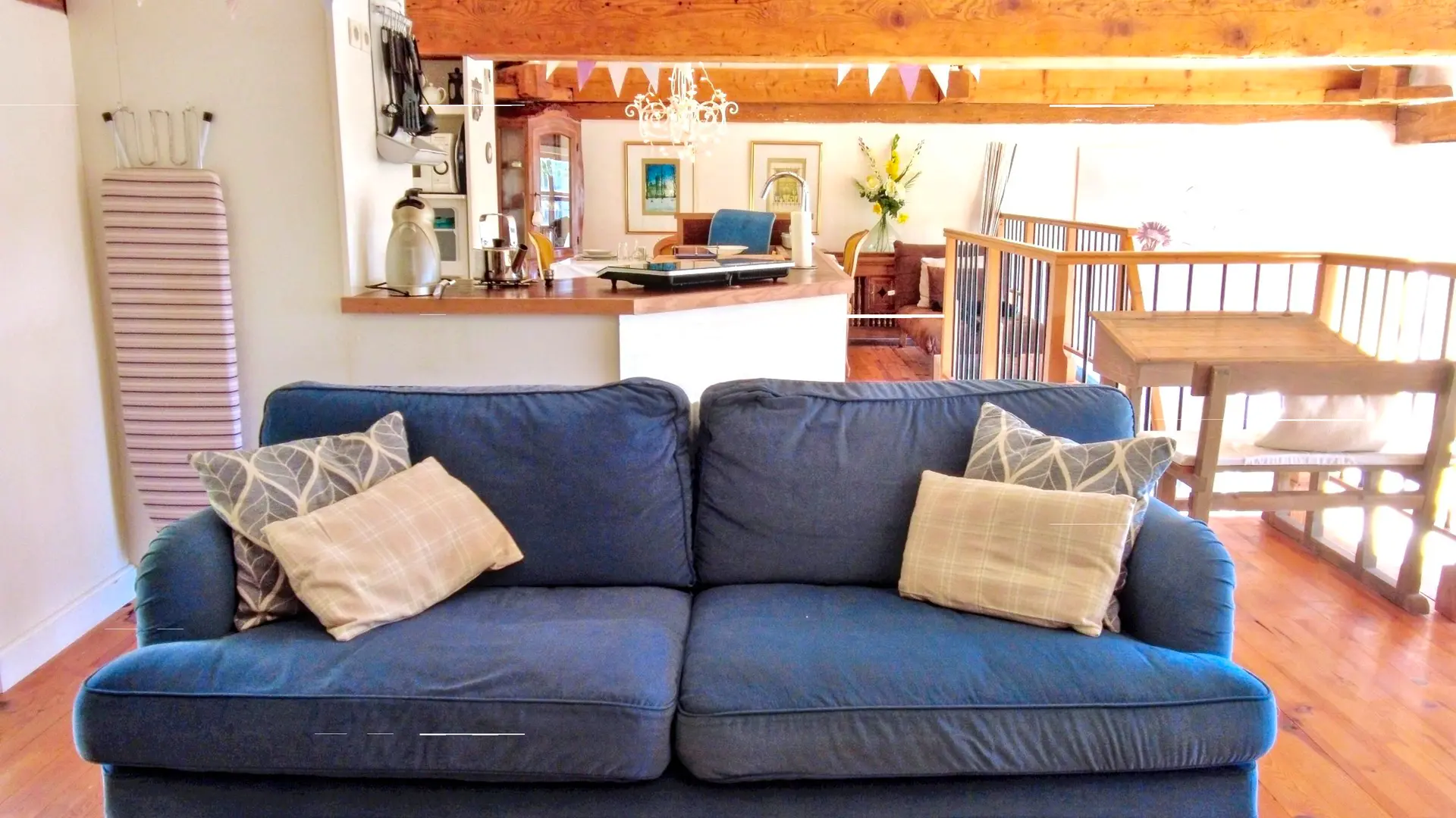Un gros plan d'un canapé deux places en tissu bleu profond, placé au centre du salon. Le canapé est garni de quatre coussins, deux à motifs géométriques gris et blancs, et deux de couleur beige à carreaux. La pièce a un plancher en bois et un plafond avec des poutres en bois apparentes. À l'arrière-plan, au-dessus du canapé, on aperçoit un bar ou un comptoir de cuisine.