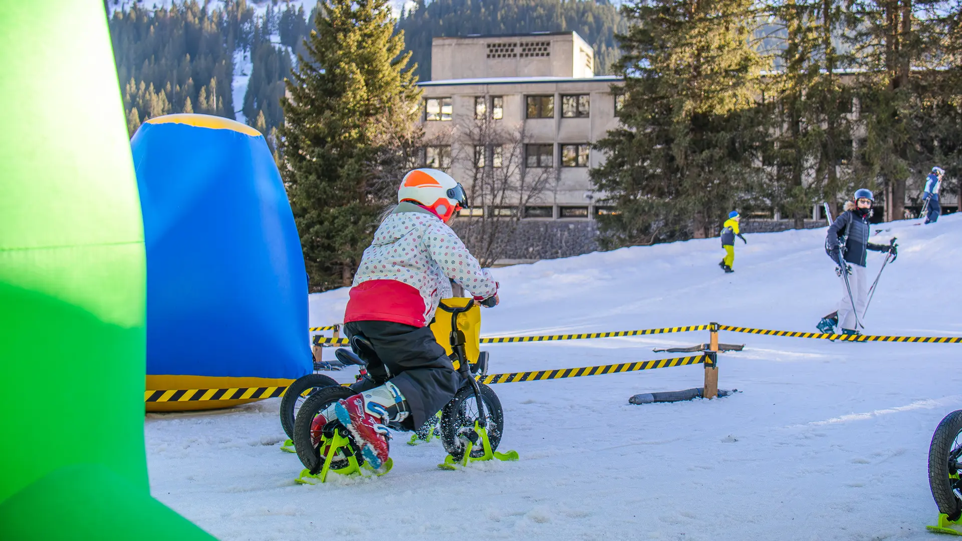 Un enfant portant un casque et une veste colorée s'entraîne sur un petit vélo-ski à l'intérieur d'un parcours balisé sur la neige.