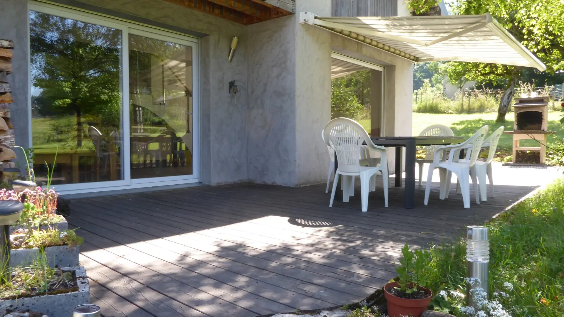 Terrasse ombragée avec table et chaises sous un store et barbecue en pierre.