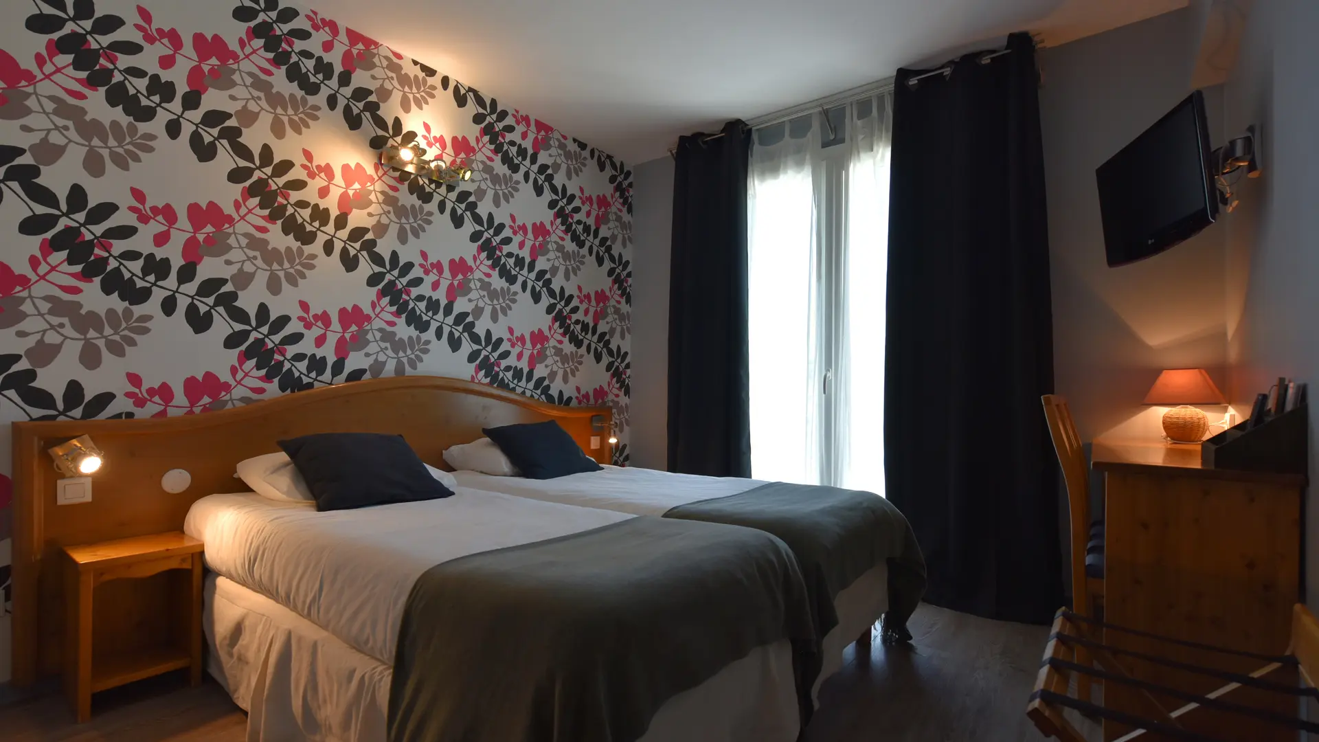 Chambre avec deux lits simples, chacun accompagné d’une table de chevet éclairée par un spot. Une applique murale et une lampe de bureau complètent l’éclairage. Présence d'un téléviseur et d'un porte bagage
