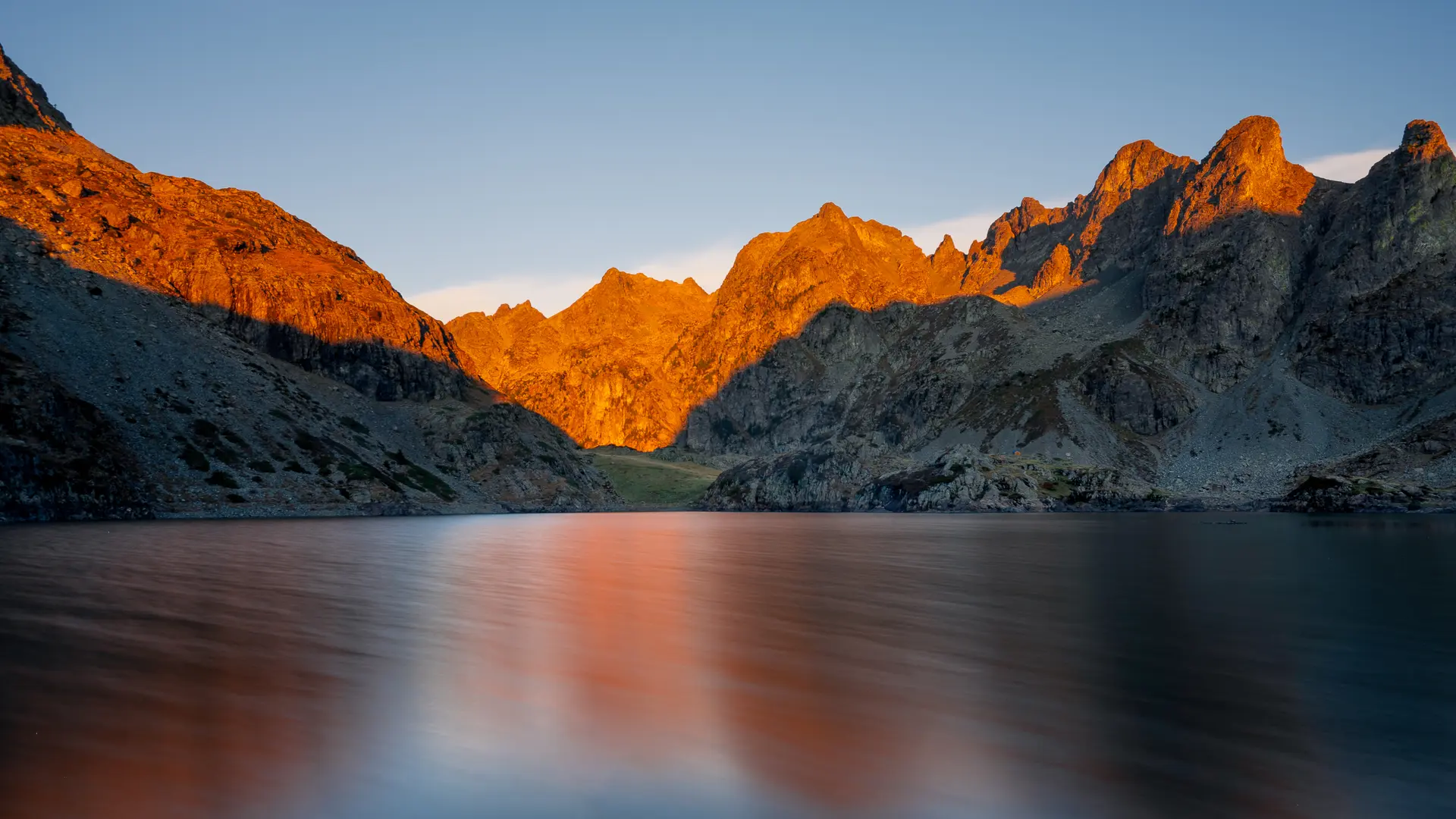 Un lac s'étend sous un ciel clair, avec des montagnes escarpées et irrégulières qui s'élèvent sur les côtés.