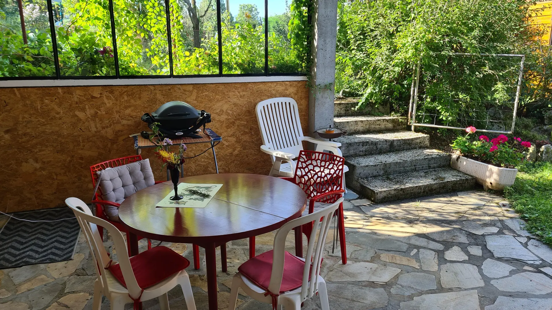 C'est une grande baie vitrée extérieure avec une grande baie vitrée donnant sur un jardin. On y trouve un barbecue, un salon de jardin composé de chaises et d'une table, ainsi que quelques objets de décoration.