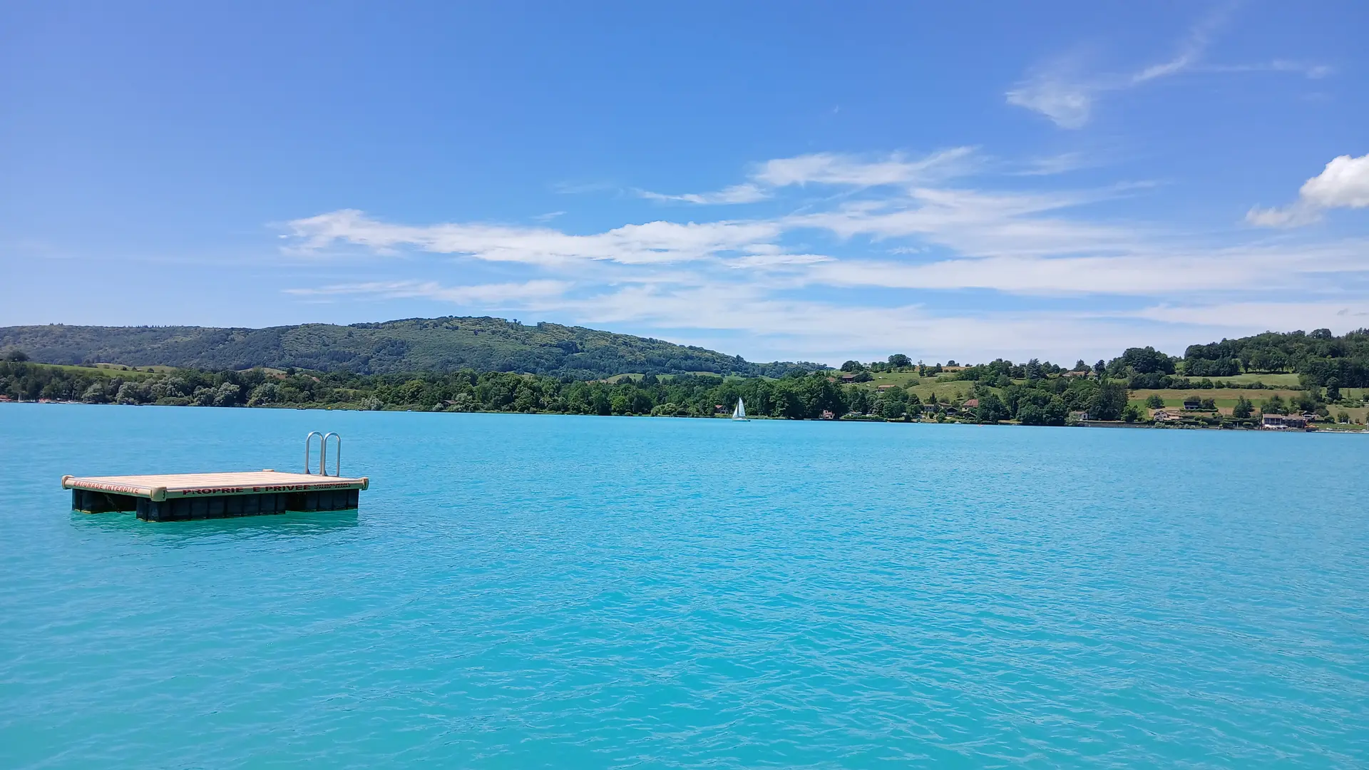 Un radeau et son échelle flottent fièrement sur les eaux cristallines d'un lac turquoise. Personne à déceler sur l'entièreté de la vue large et calme. Au fond, une planche à voile défile devant la rive opposée.