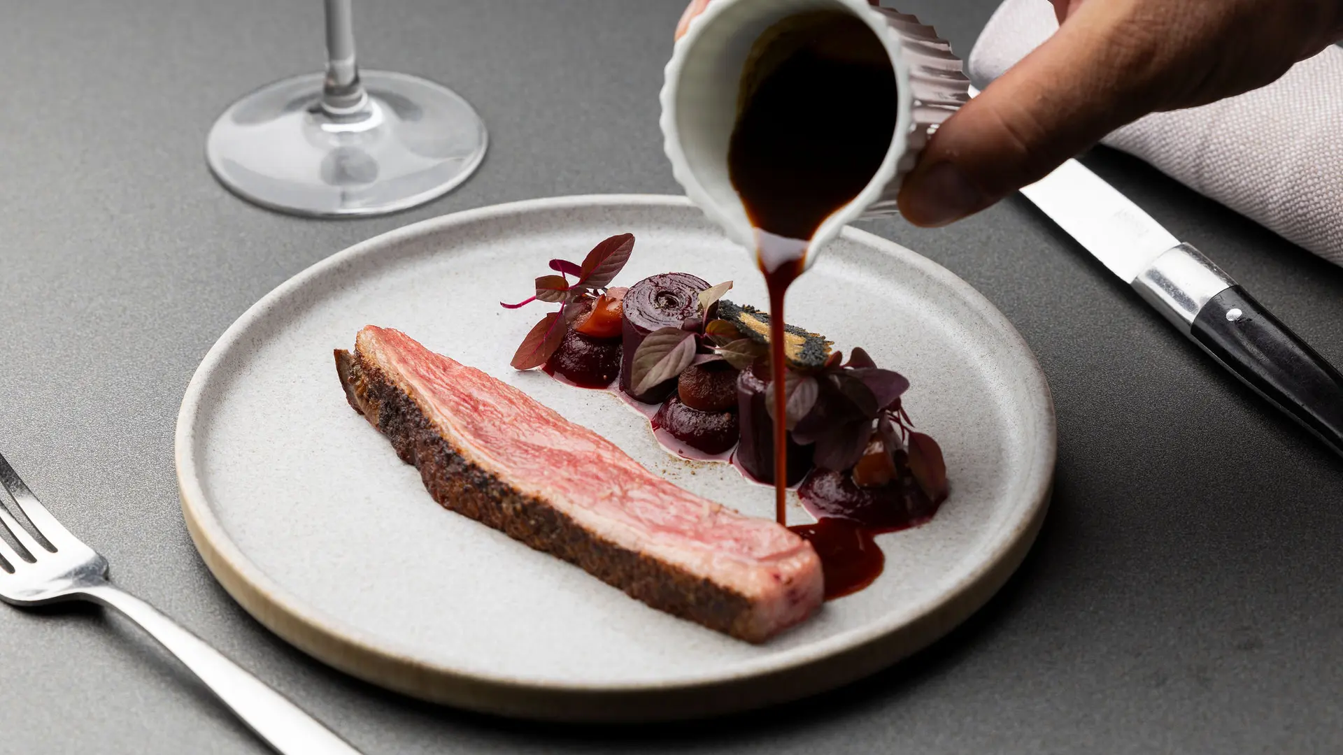 Sur une assiette ronde de couleur grise, une tranche de viande cuite est alignée avec une garniture de légumes ronds et de feuilles pourpres, tandis qu'une main verse une sauce foncée dessus à partir d'une petite saucière cannelée.