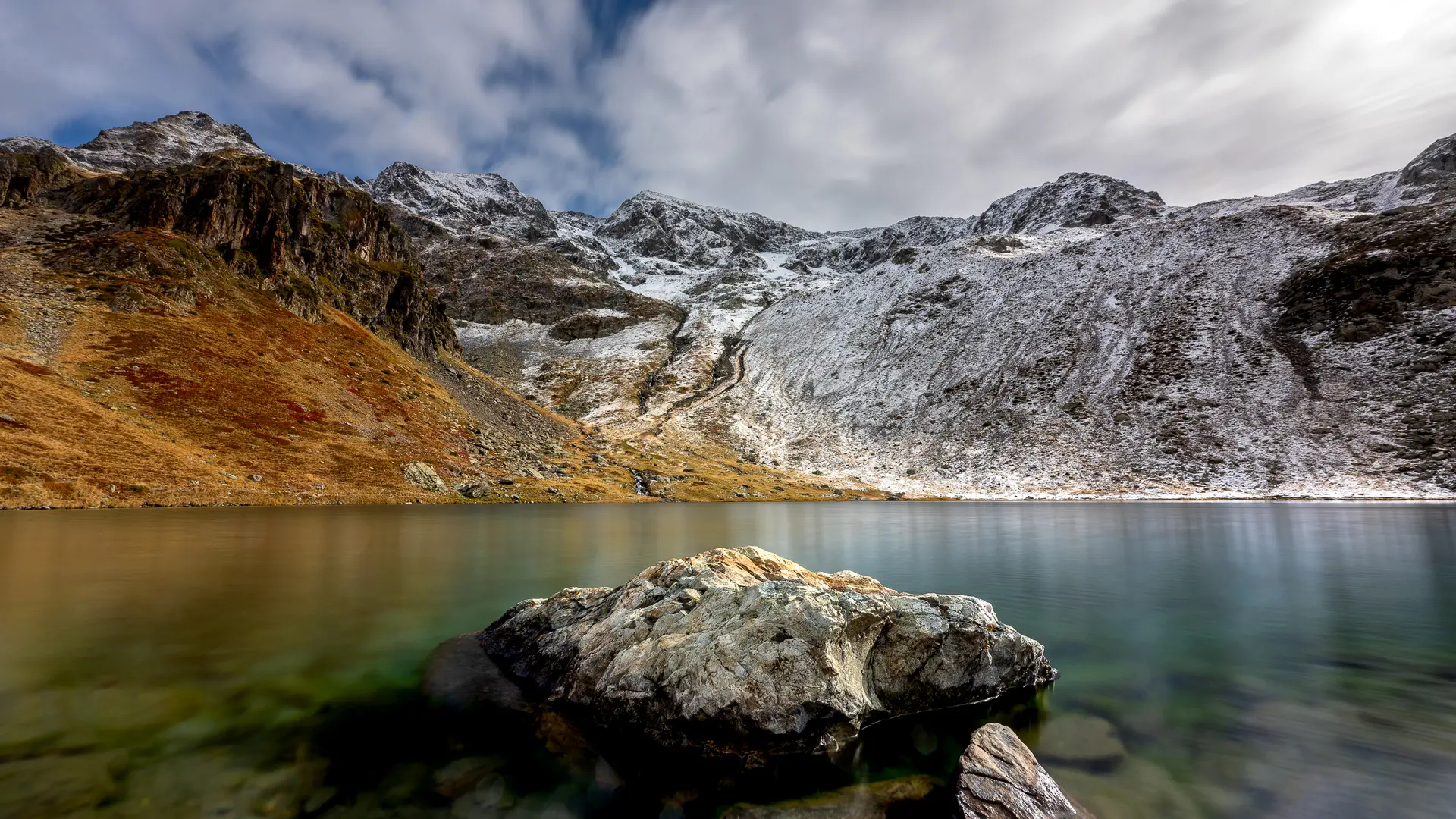 Un lac s'étend sous un ciel nuageux, avec des montagnes escarpées et irrégulières qui l'entourent.