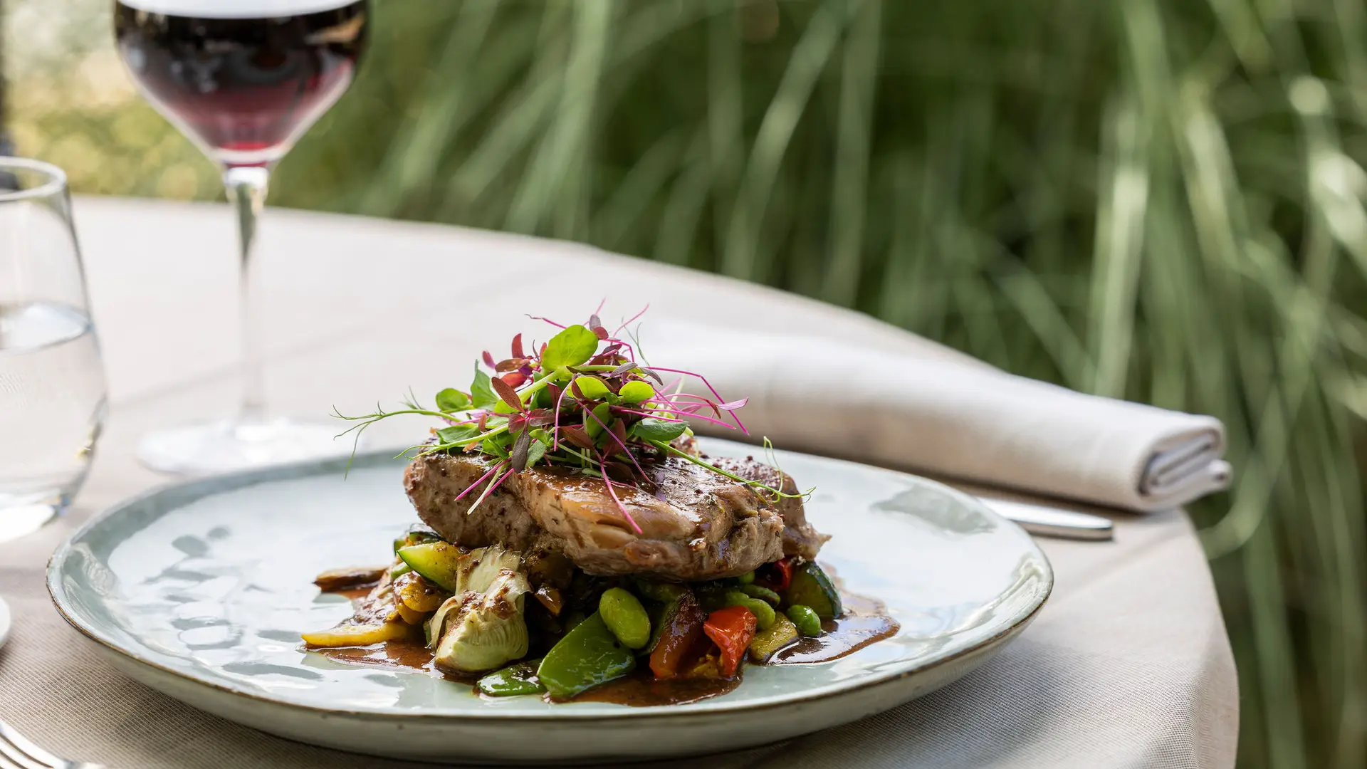 Sur une assiette ronde vert pâle et entourée d'une table avec un verre de vin et un verre d'eau en arrière-plan flou, une portion de viande rôtie repose sur un lit de légumes sautés recouverts d'une sauce, le tout garni d'une touffe de jeunes pousses.