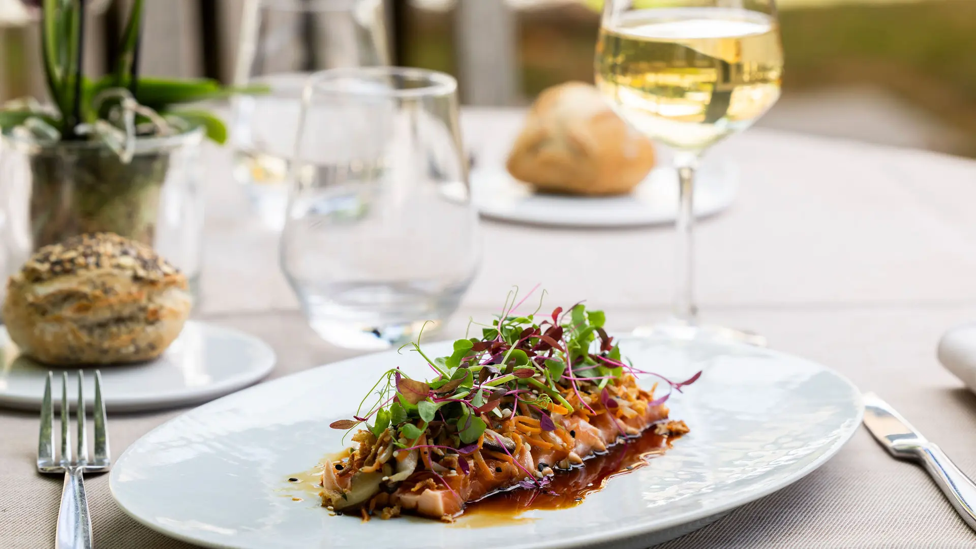 Sur une table dressée avec un verre de vin blanc et de l'eau, un plat est présenté sur une assiette ovale blanche.