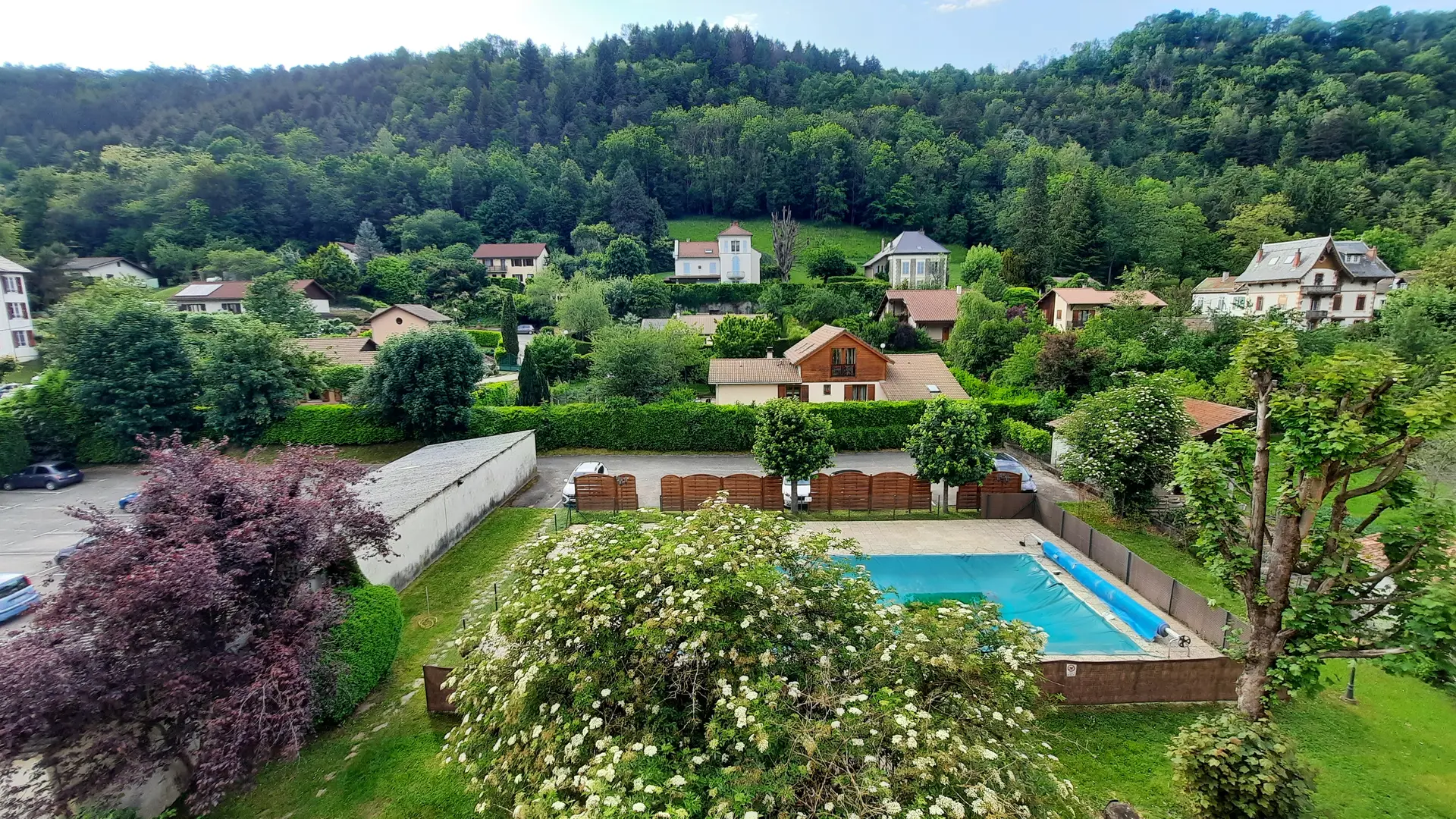 Un paysage paisible avec une piscine au premier plan, entourée de verdure. Au fond, des maisons sont dispersées sur les collines boisées.