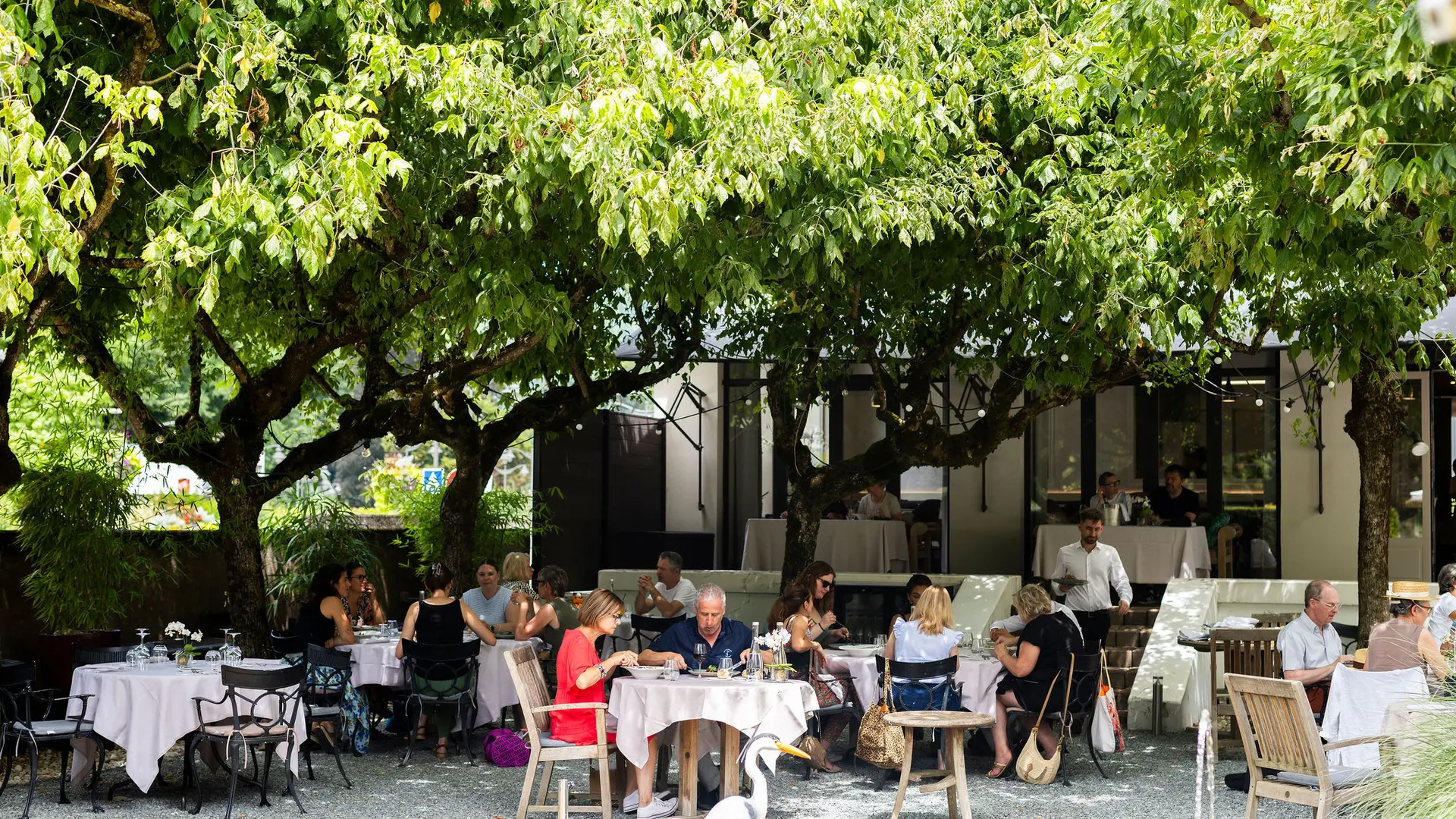 Une terrasse de restaurant ombragée par de grands arbres au feuillage dense, où plusieurs tables recouvertes de nappes claires sont dressées et occupées par des clients mangeant en extérieur, avec un héron décoratif au premier plan près d'un bassin.