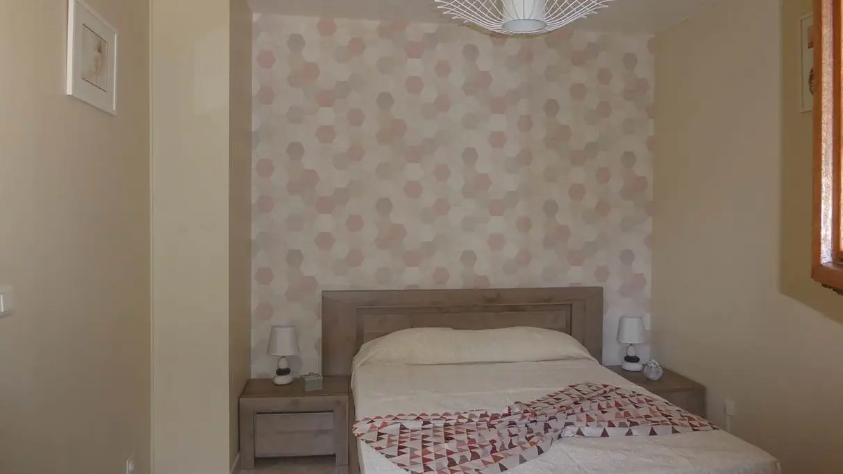 L'image montre une chambre à coucher lumineuse avec un grand lit habillé d'une couette blanche et d'un plaid à carreaux. Une fenêtre laisse entrevoir un extérieur.