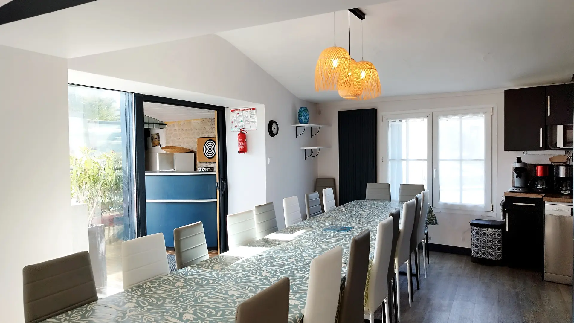 Salle à manger avec porte-fenêtre ouverte sur l'extérieure, tables et chaises pour une quinzaine de personnes.