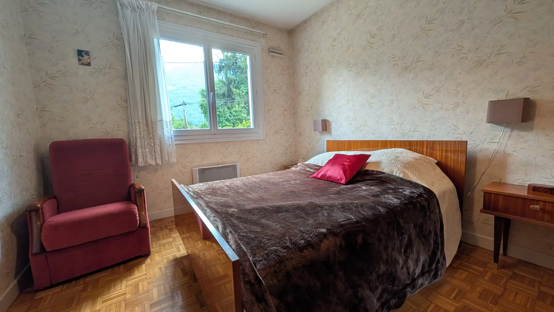 Cette chambre à coucher lumineuse comprend un lit double, flanqué à gauche d'un fauteuil bordeaux et d'une fenêtre donnant sur le jardin, et à droite d'un petit chevet en bois.