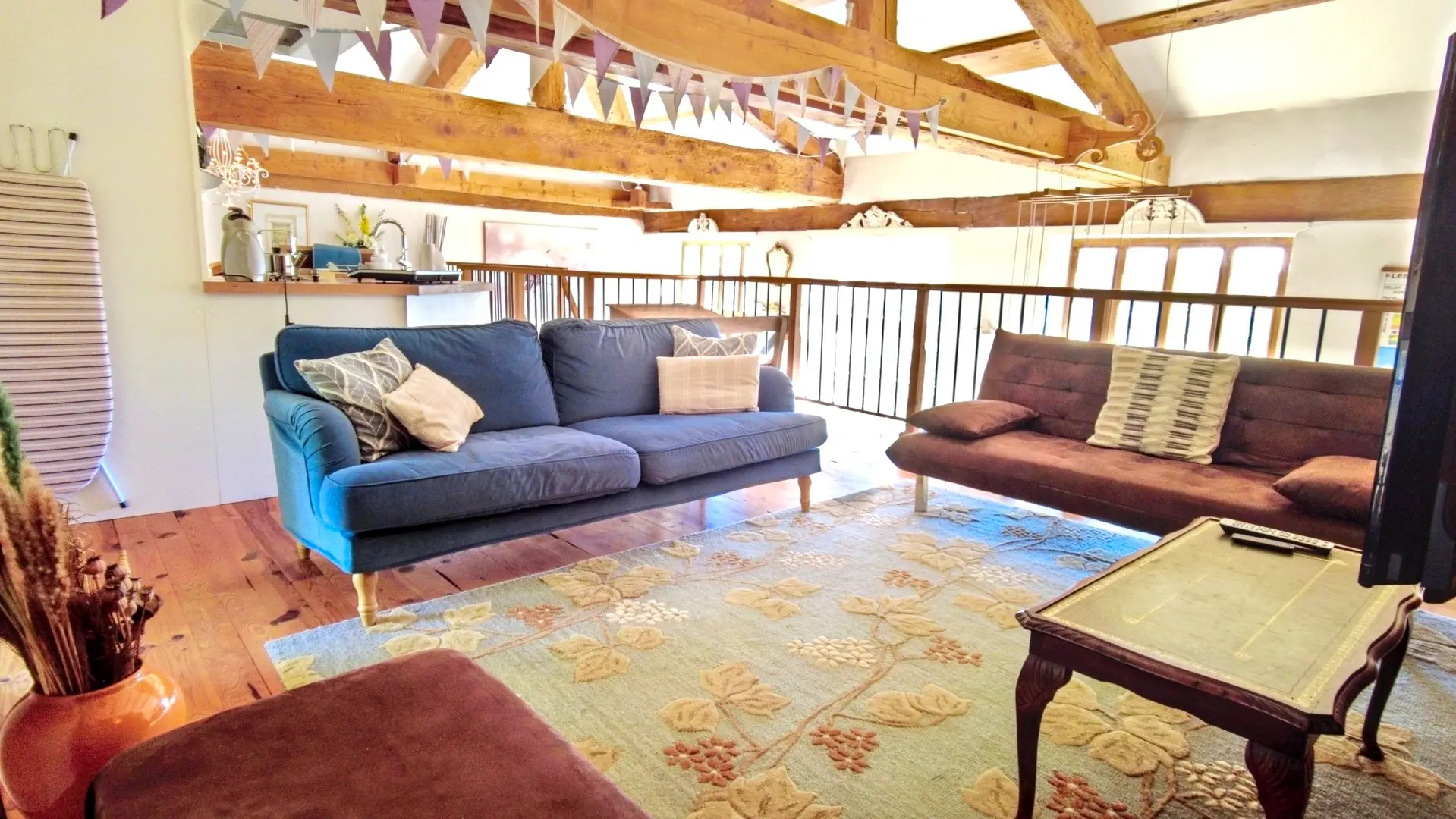 Nous sommes dans le salon de la mezzanine. Il comprend deux canapés : un bleu avec des coussins et un marron. Le plancher est recouvert d'un tapis. Une table basse se trouve en dessous de la télé. Du salon, on a une vue de la mezzanine avec sa rambarde.