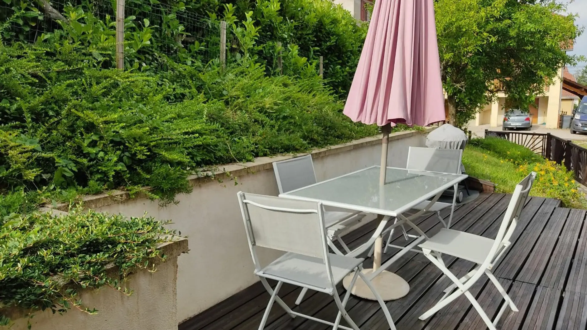 Sur une large terrasse au pied d'une haie verdoyante, une longue table de verre et ses quatre chaises accueillent un parasol pour créer une zone propice aux repas extérieurs. Derrière, un petit barbecue est protégé par une bâche.