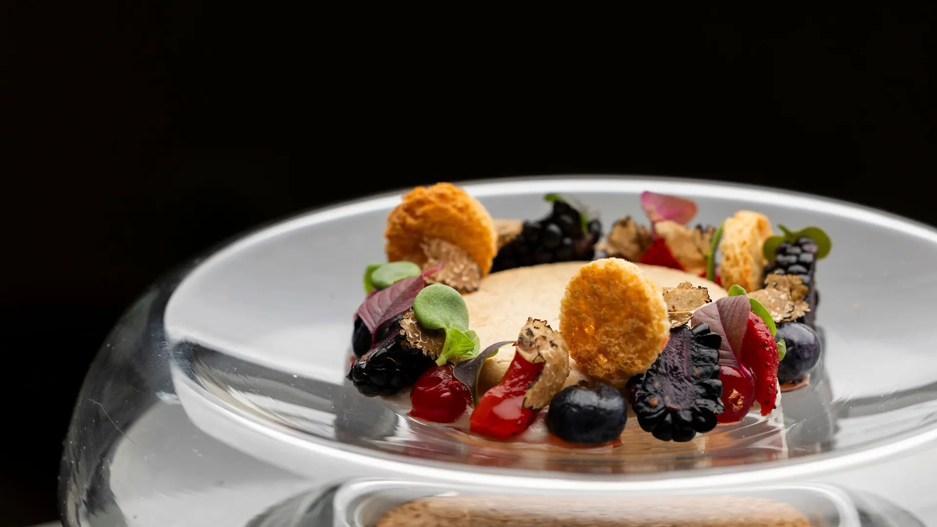 Sur un socle circulaire transparent, une petite assiette en verre contient un dessert en forme de couronne, composé d'une base claire recouverte d'un anneau de fruits des bois noirs et rouges, de petites tuiles rondes dorées et de quelques feuilles vertes.